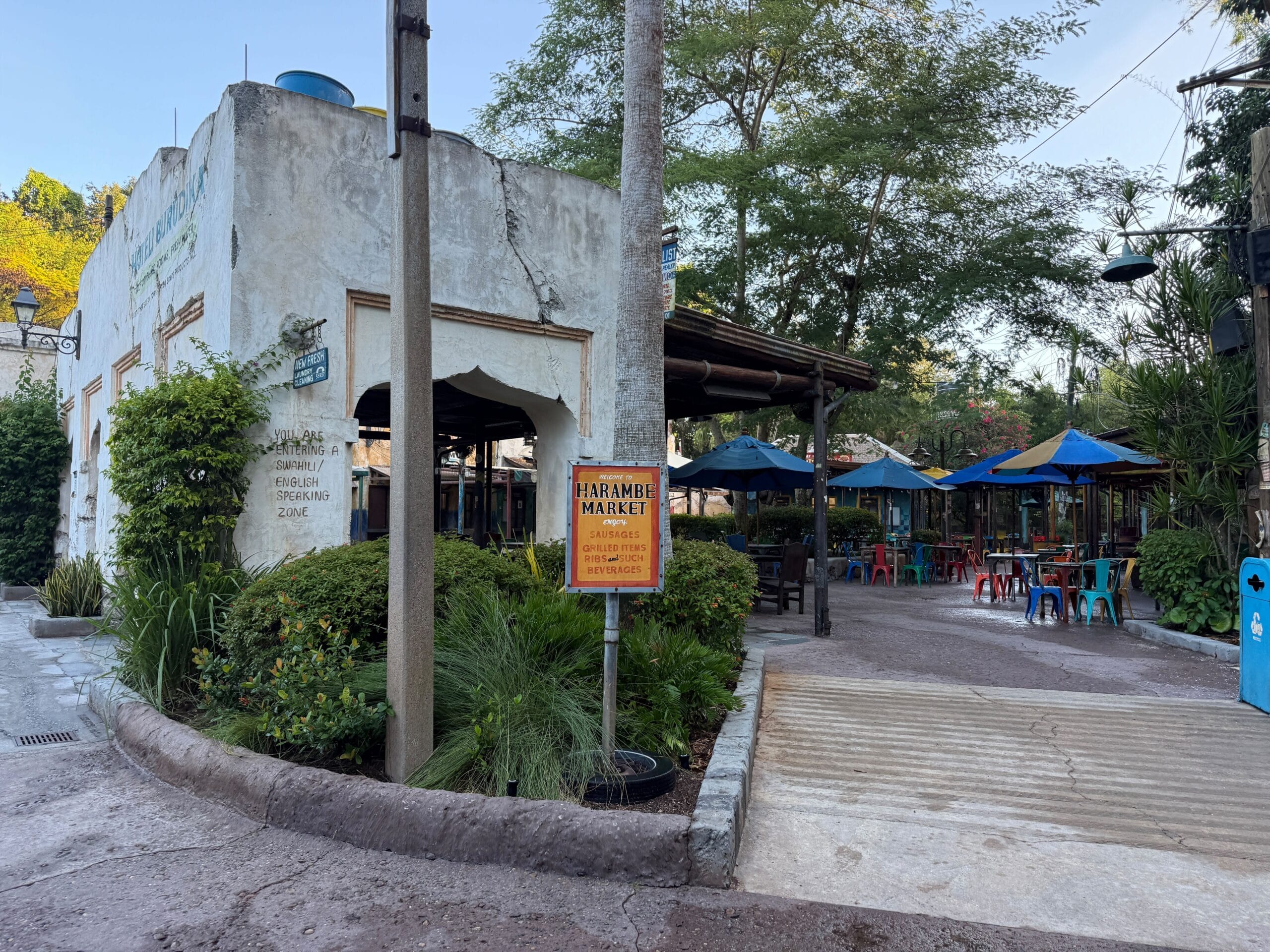 Harambe Market’s outdoor seating in Disney’s Animal Kingdom with lush greenery, shaded umbrellas, and a friendly sign—no castle nearby.
