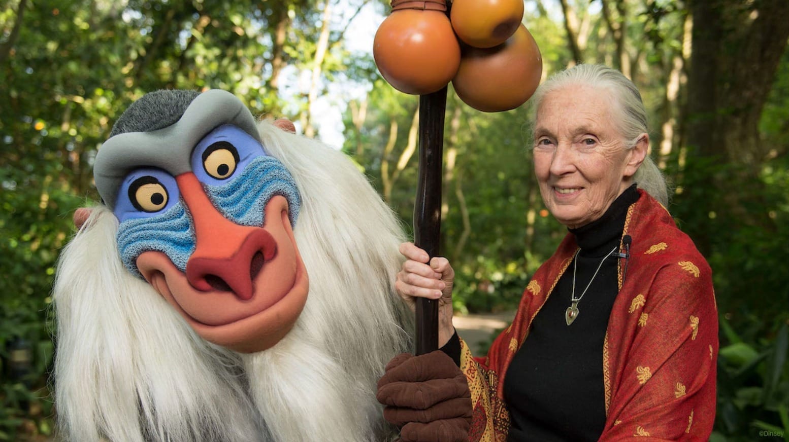 A woman stands with a person in a colorful baboon costume, honoring Jane Goodall in a lush, green outdoor setting.