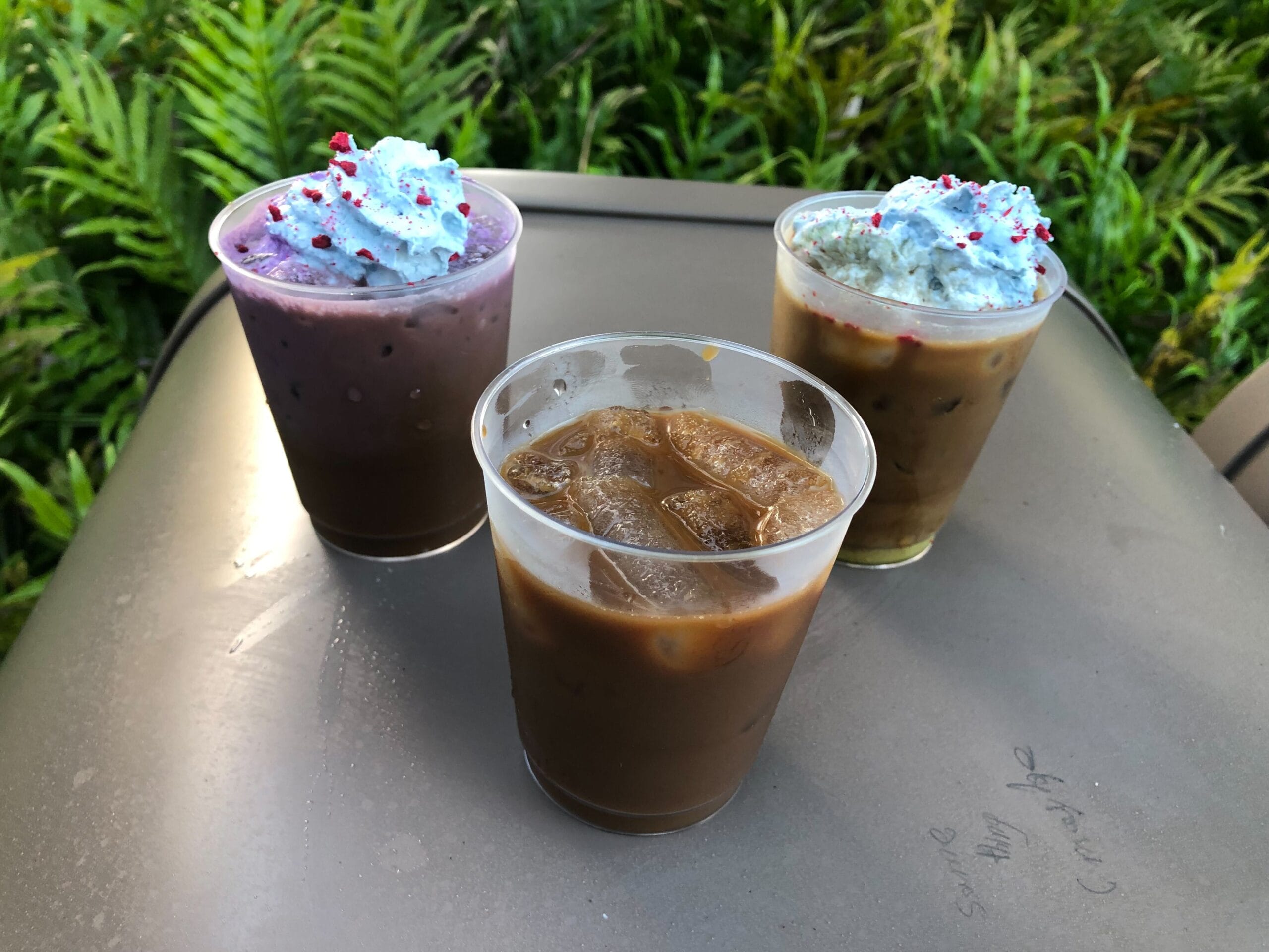 Three iced coffee drinks on a table, two with whipped cream and sprinkles, with green plants in the background.