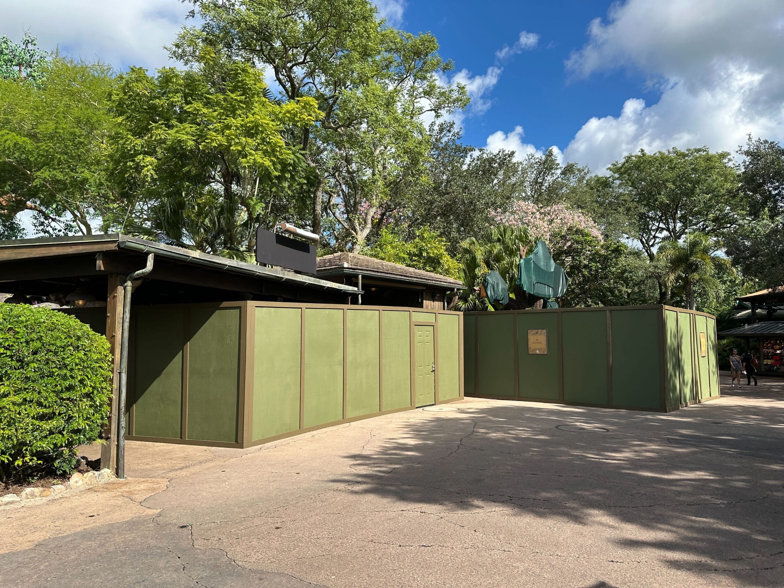 Green construction walls enclose a section in Animal Kingdom, with lush trees and a partly cloudy sky typical of Walt Disney World.