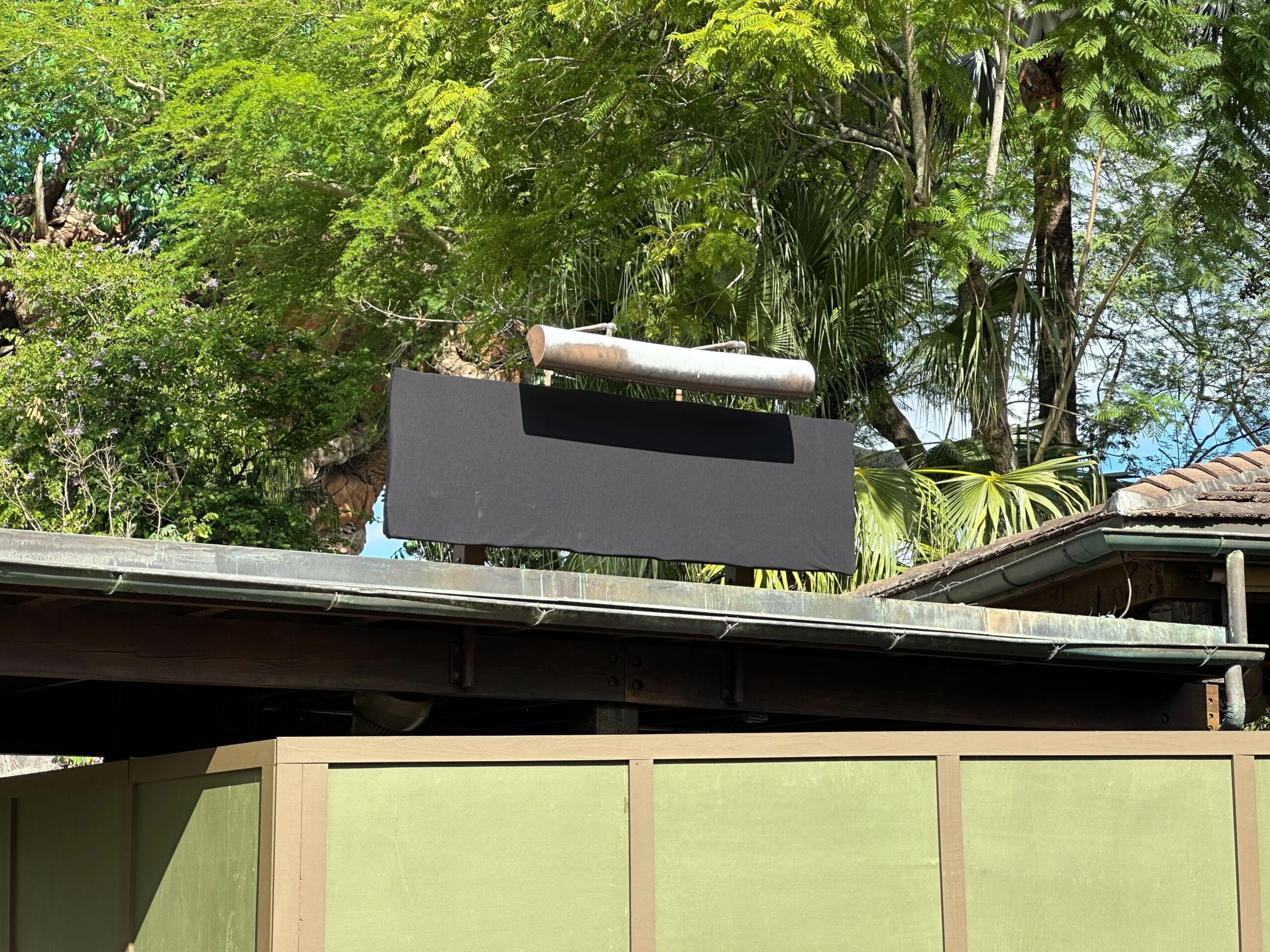 A blank black Animal Kingdom Construction sign sits atop a roof, surrounded by lush greenery, evoking Disney’s natural park setting.