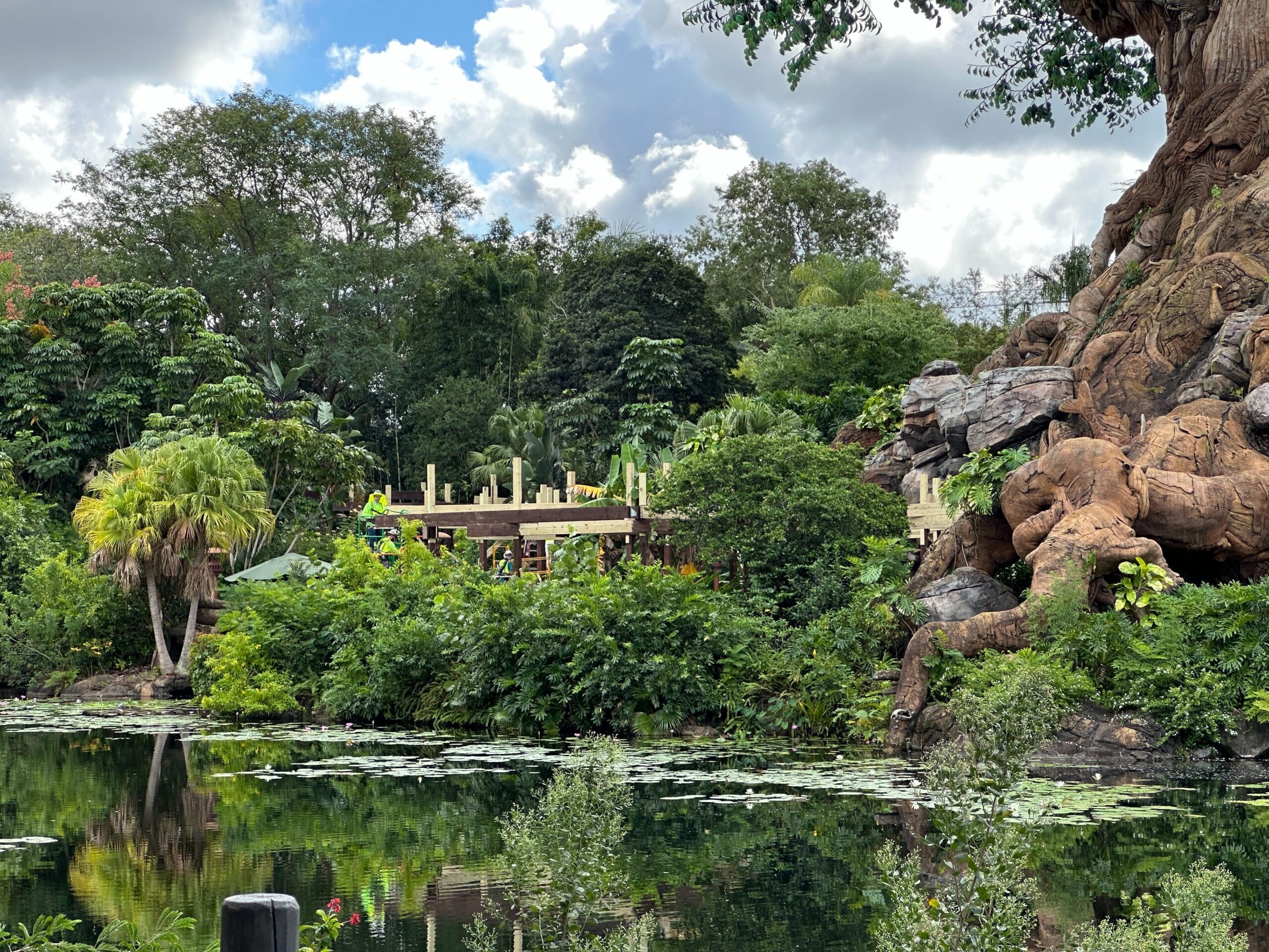 A vibrant Animal Kingdom-style scene with tropical plants and a pond under partly cloudy skies, reminiscent of Disney park landscaping.