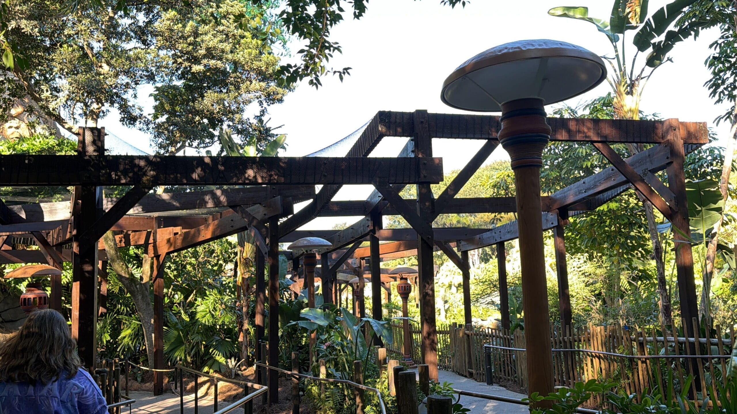 A winding outdoor walkway with wooden pergolas, heaters, and lush greenery, reminiscent of a Disney park garden, sunlit with a guest ahead.
