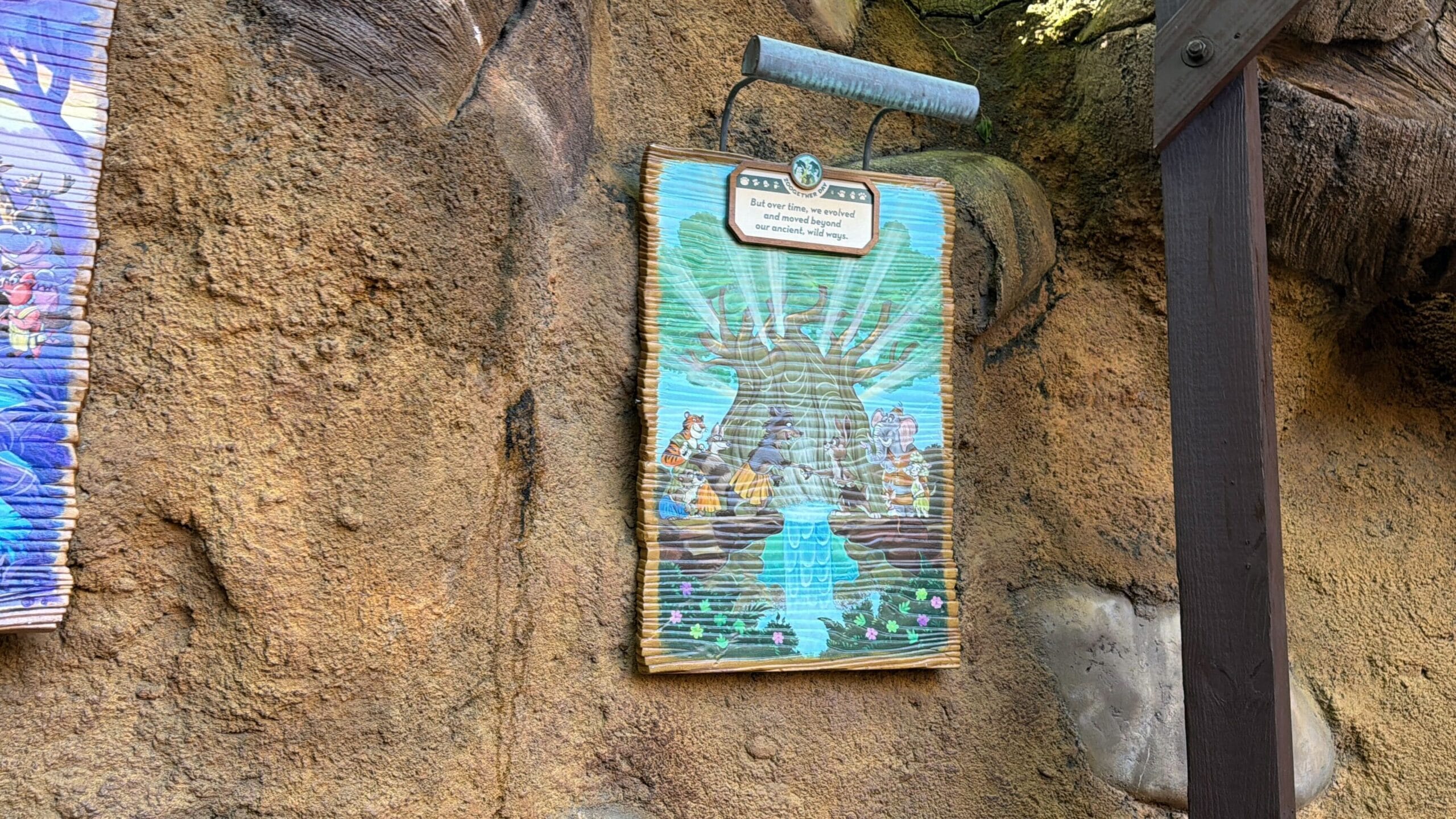 A vibrant sign featuring cartoon animals and a tree decorates a rocky wall, reminiscent of playful Disney park theming nearby shade.