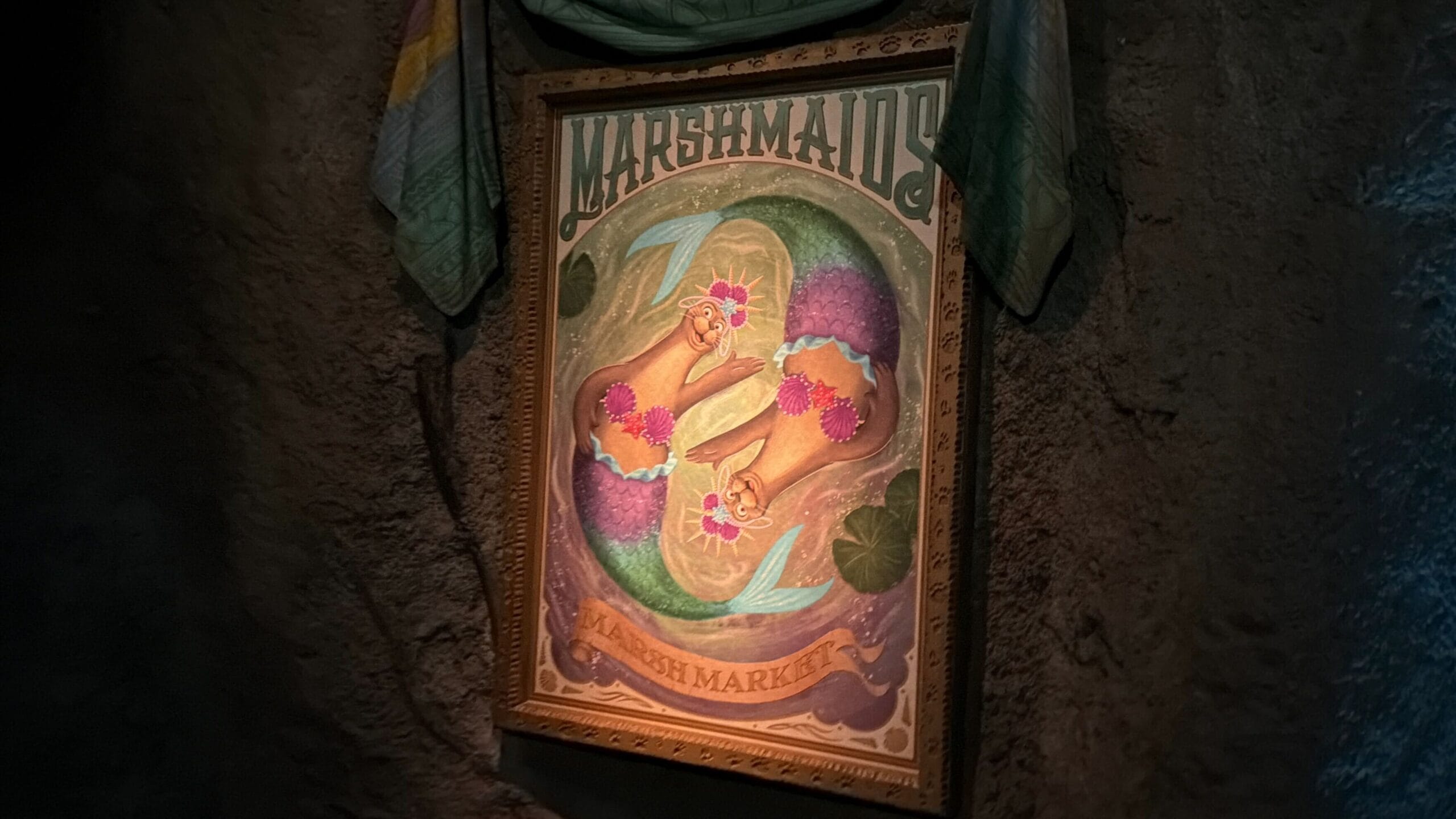 Framed poster of two illustrated mermaids with flowers, “Marshmaids Marsh Market,” on a stone wall—no Disney castle visible.