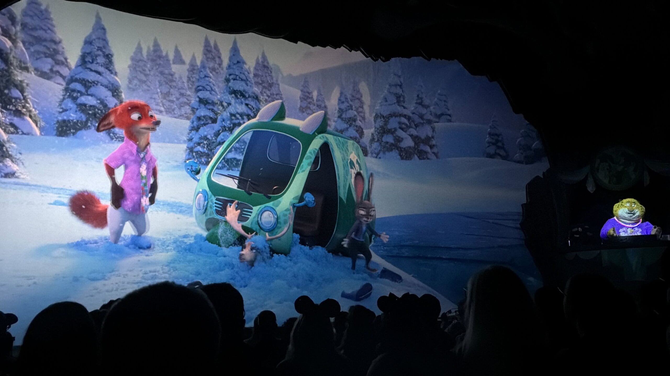 Animated scene with animals near a green van in snowy surroundings, viewed by guests in a dark Disney park theater, no castle visible.