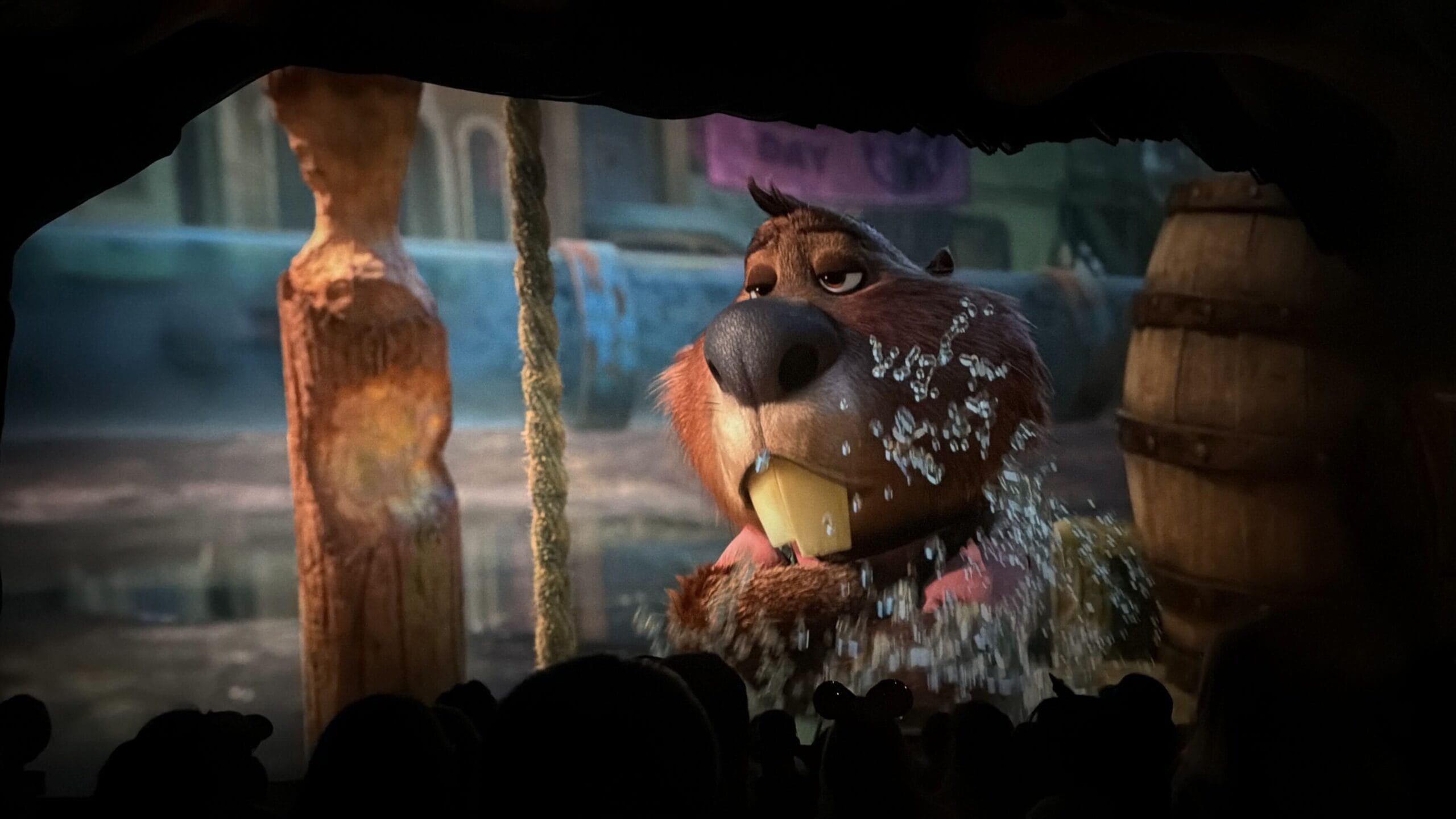 Animated otter-like character playfully splashes water on stage, viewed from a dark Disney park theater with audience silhouettes.