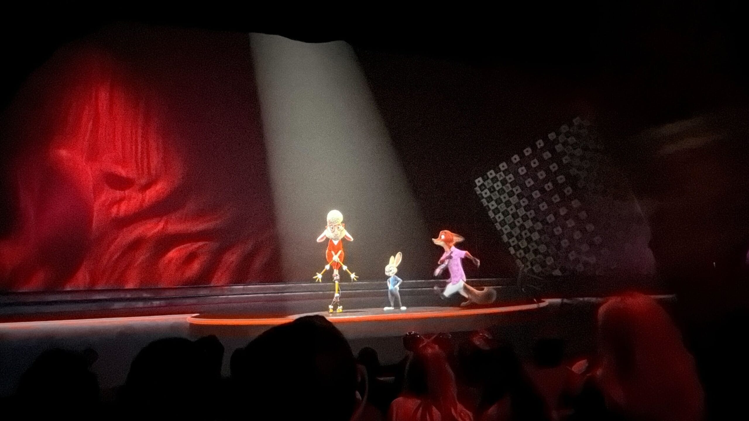 Three animated characters perform on stage under a spotlight, red and black backdrop behind, audience silhouettes like Disney park theaters.