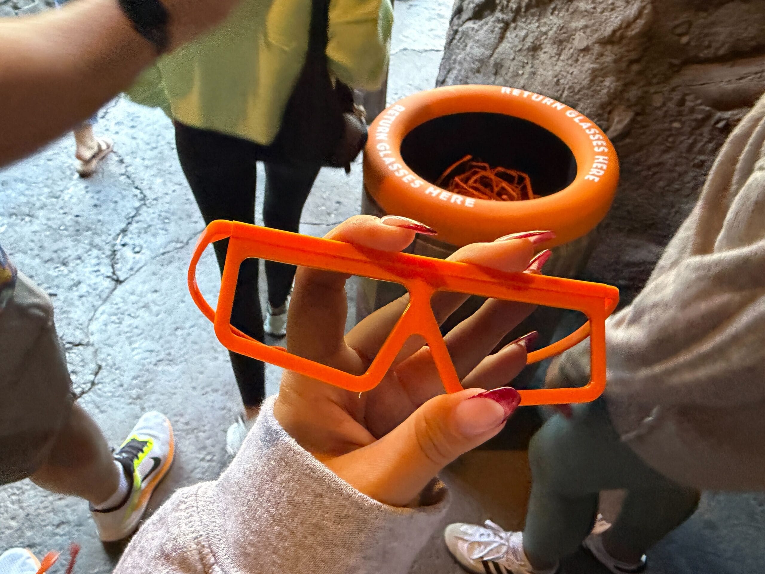 A hand holds orange 3D glasses near a "Return Glasses Here" bin at Animal Kingdom, Disney World.