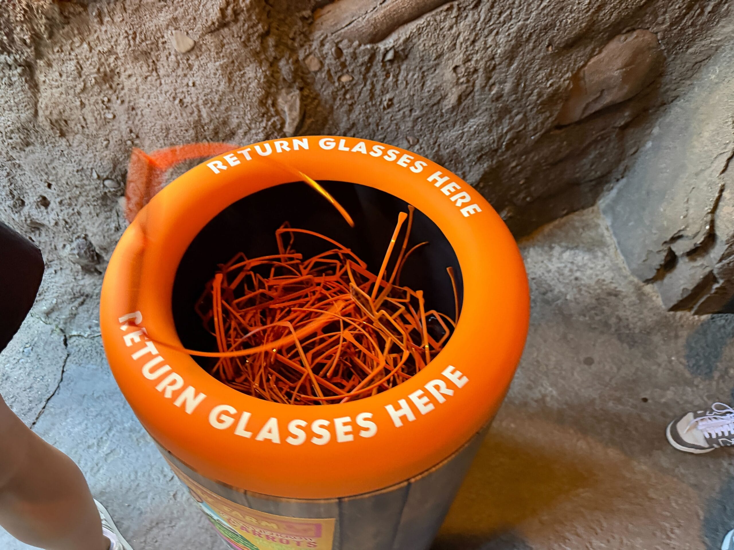 Orange bin labeled "Return Glasses Here," filled with orange 3D glasses frames, spotted near a Disney park attraction entrance.