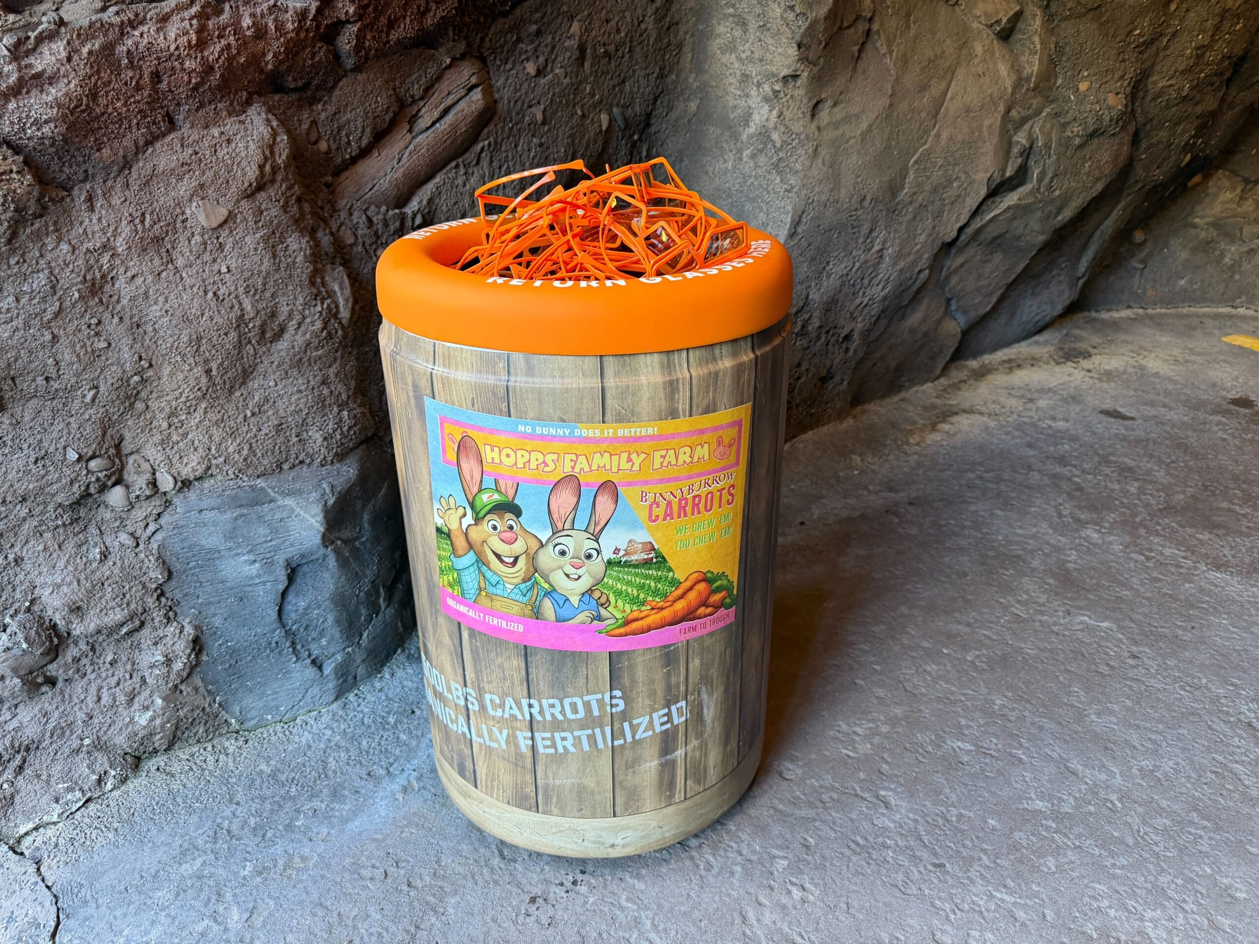 A wooden barrel-shaped trash can at a Disney park, featuring cartoon rabbits, "Hopps Family Farm" text, and topped with faux carrots.