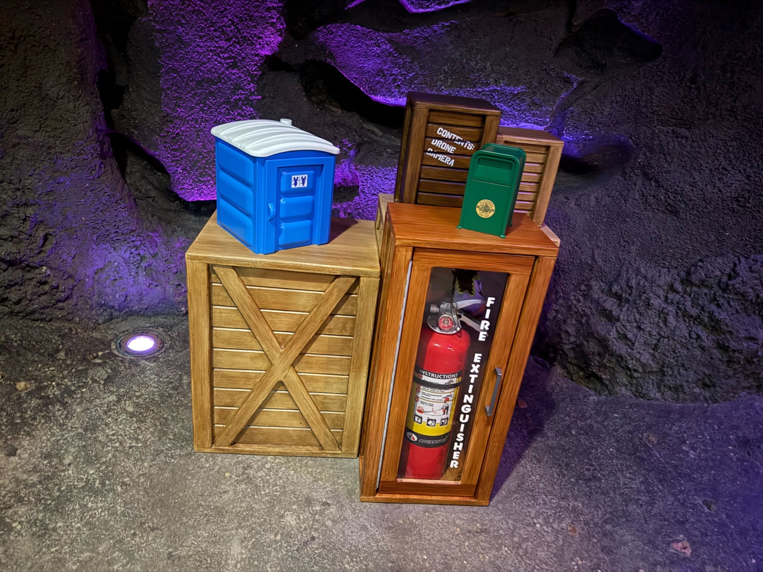 Four Disney park-themed safety props, such as a fire extinguisher, on rocks with purple lighting, evoking a ride queue atmosphere.