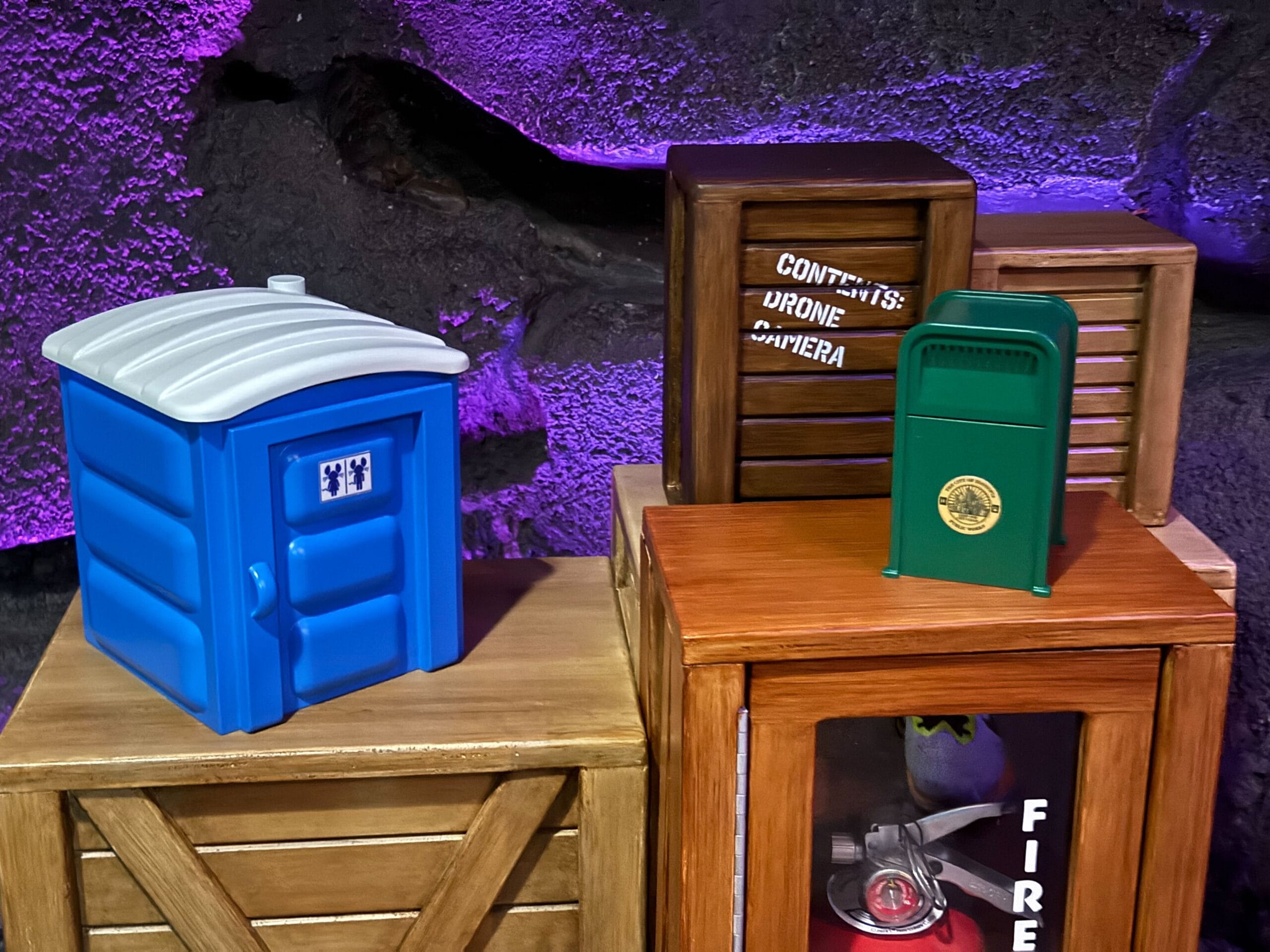 Miniature blue portable toilet, green mailbox, and wooden crates set in a Disney park-style rocky, purple-lit scene.