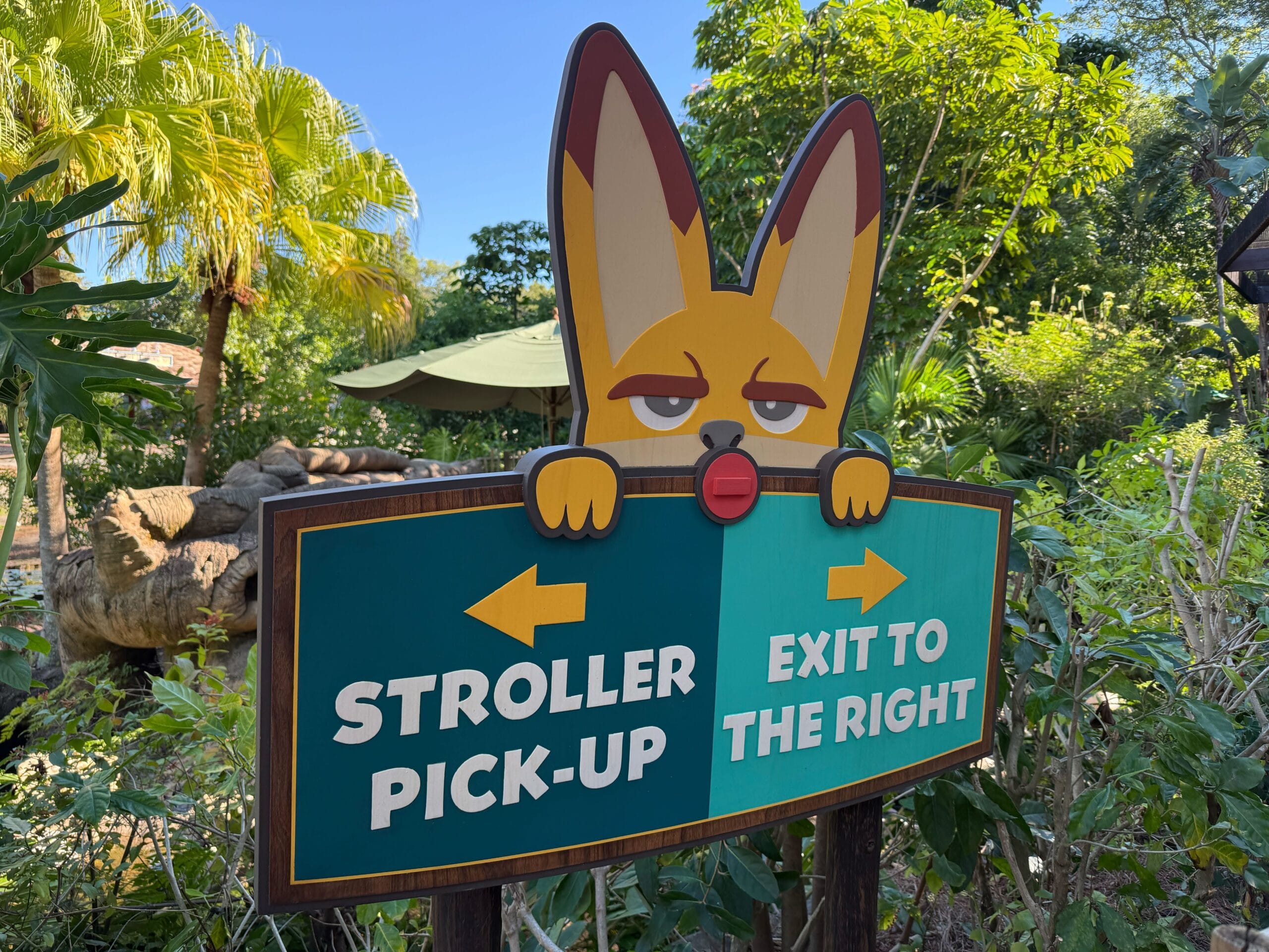 A playful cartoon animal at Disney peeks over a sign directing guests left for stroller pick-up and right for the exit.