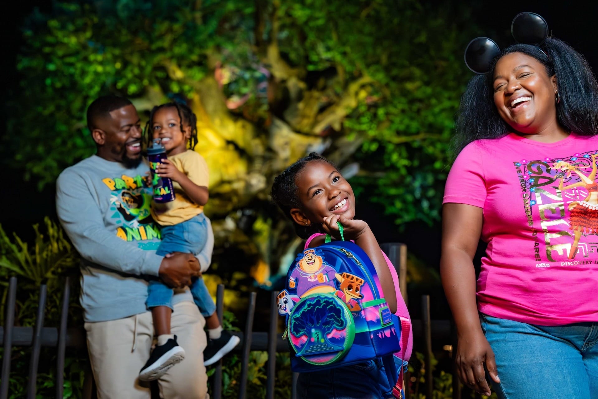 A smiling family of four stands outside at night in Disney Park; kids in bright outfits, mom sporting Mickey ears, castle not visible.