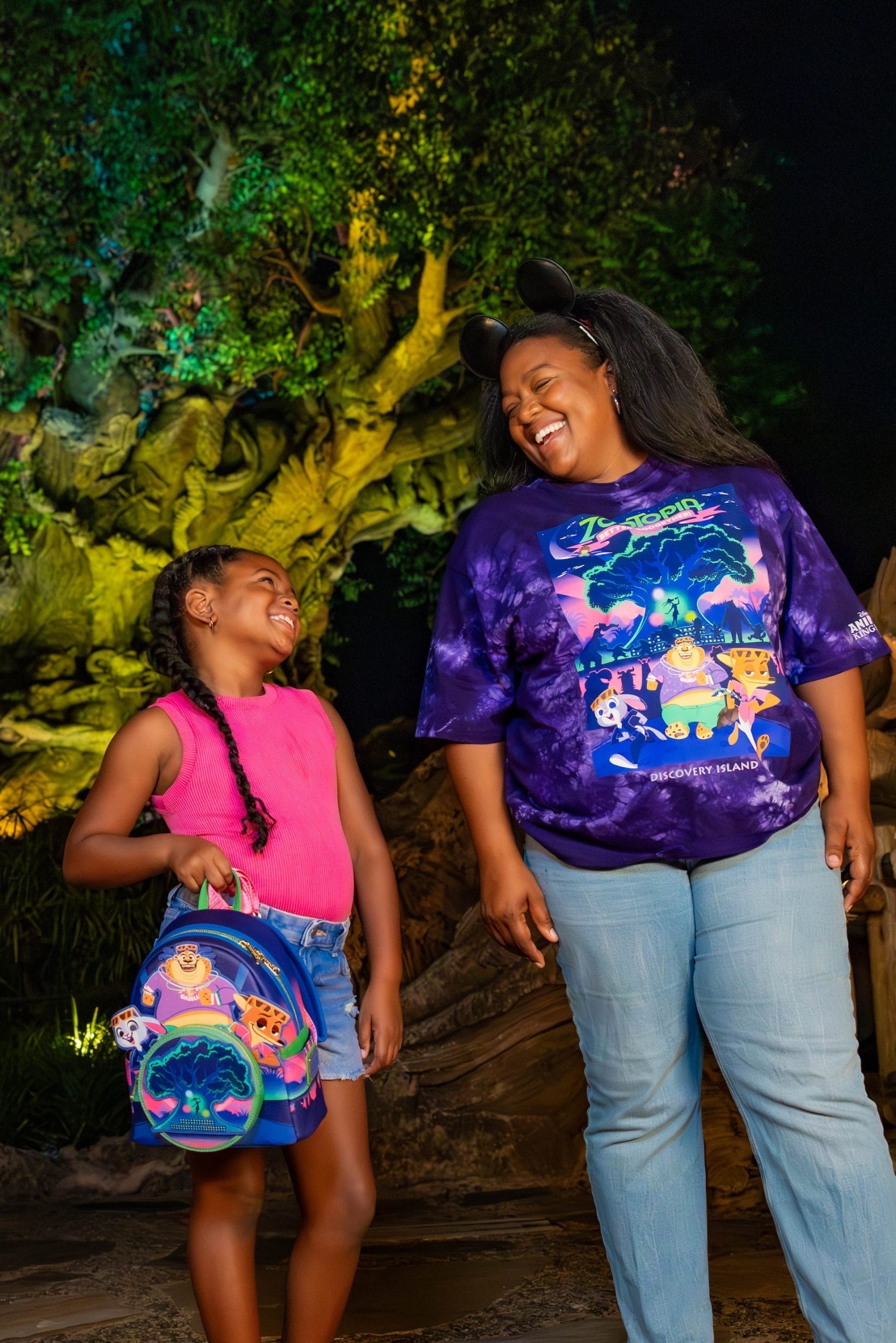 A woman and a girl share a smile in front of a large, glowing tree at night, reminiscent of Pandora’s Tree of Life in Disney parks.
