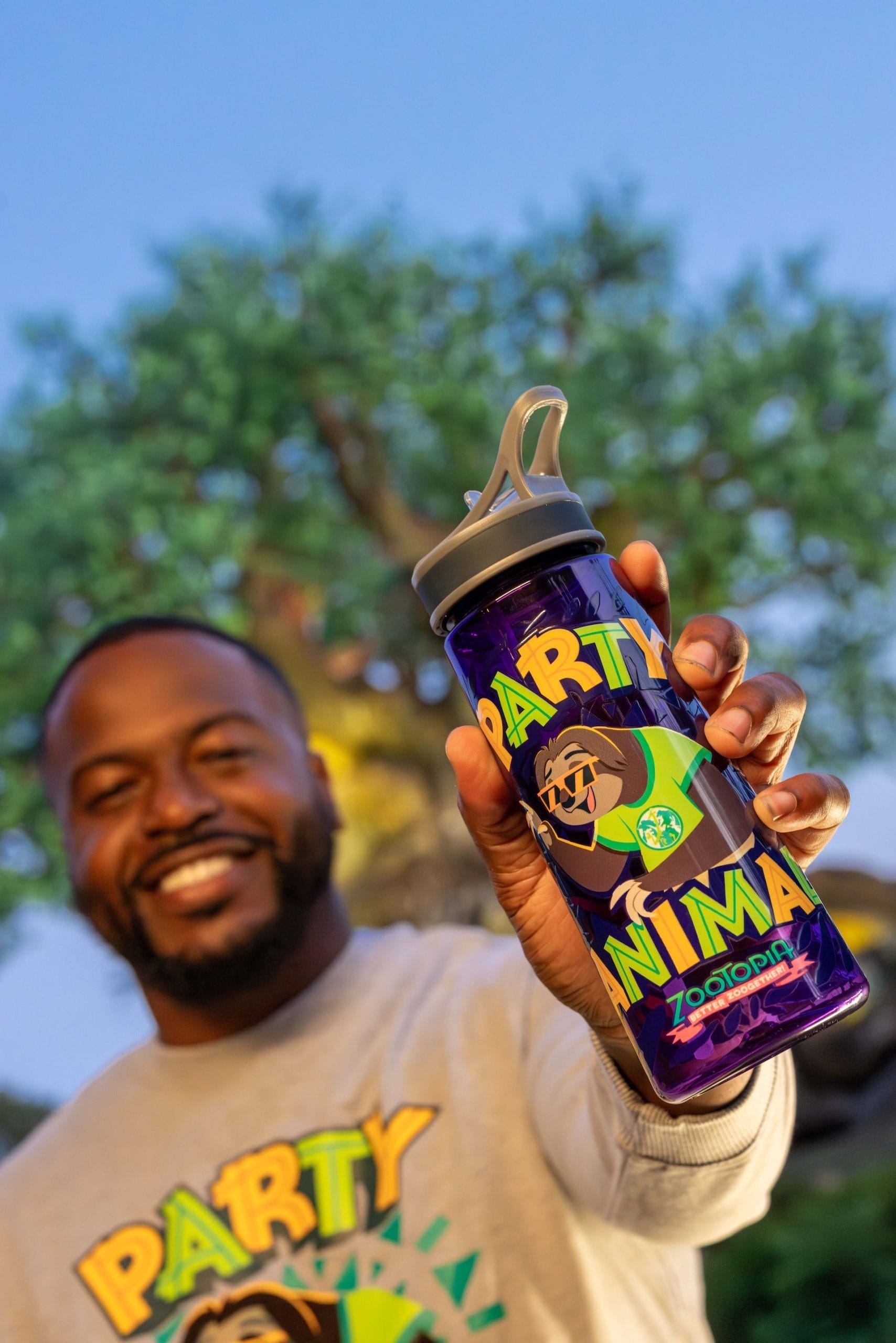 Smiling man at a Disney park holds a Zootopia "PARTY ANIMAL" bottle, standing by a tree under the clear blue sky.