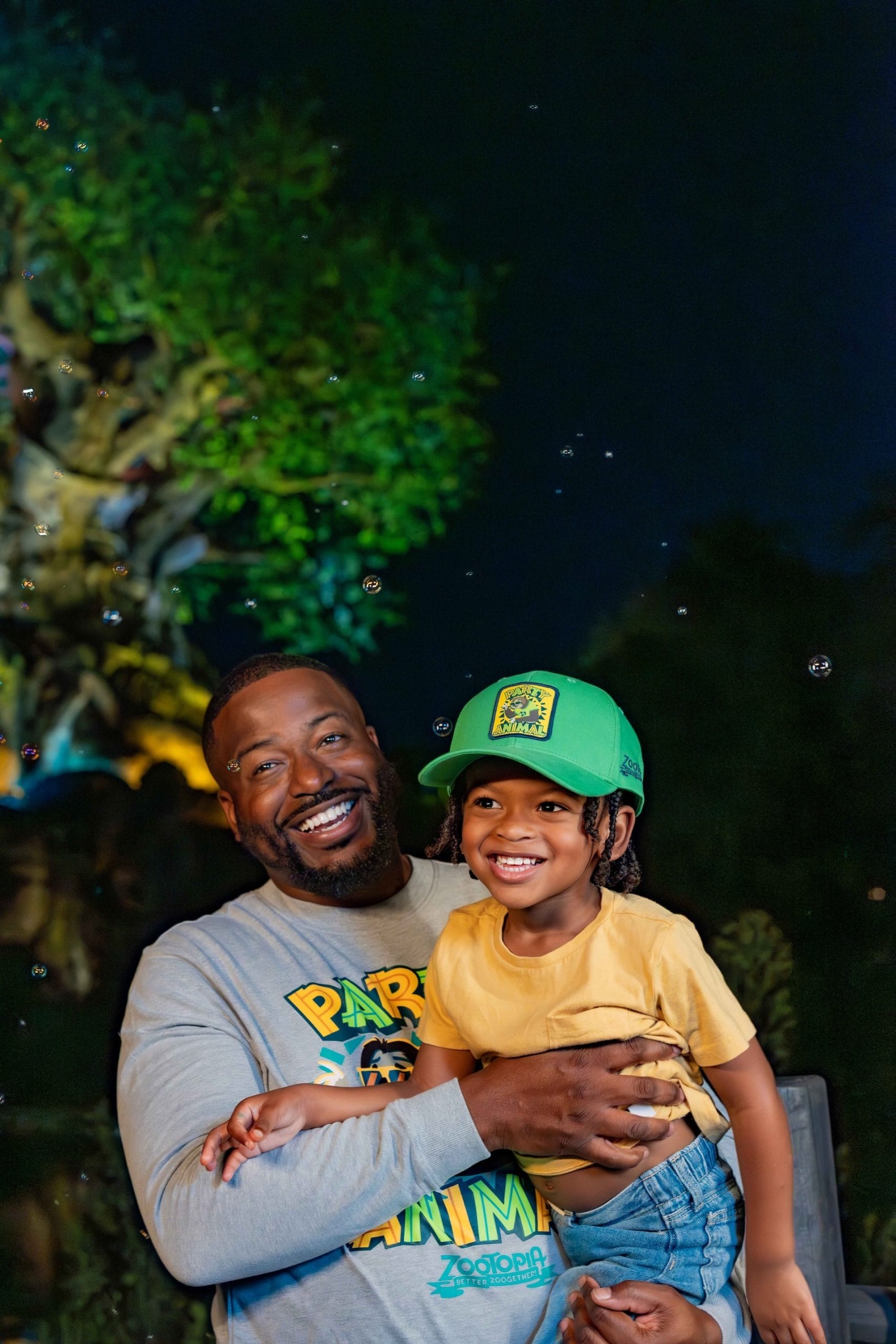 A smiling man holds a happy child outdoors at night near a sparkling tree, similar to those at Disney parks during the holidays.