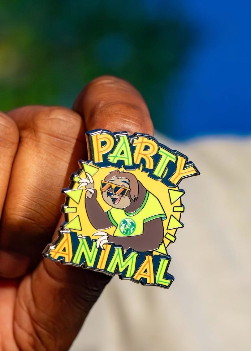 A hand displays a playful pin of a sloth in sunglasses and a shirt, reading "Party Animal," reminiscent of fun Disney park flair.