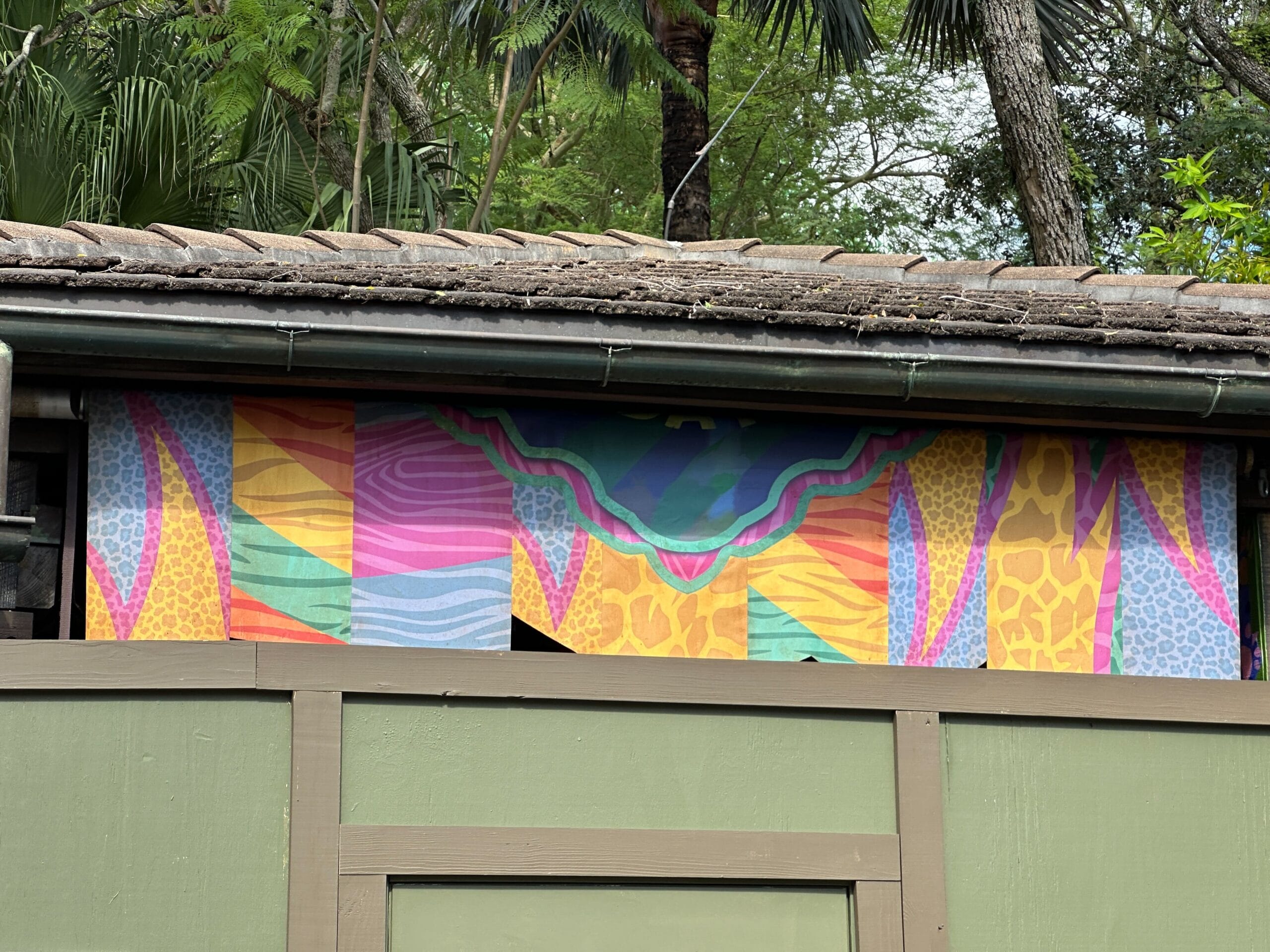 A vibrant, abstract mural with animal print patterns sits above a green fence beneath a tile roof, echoing Disney park safari themes.