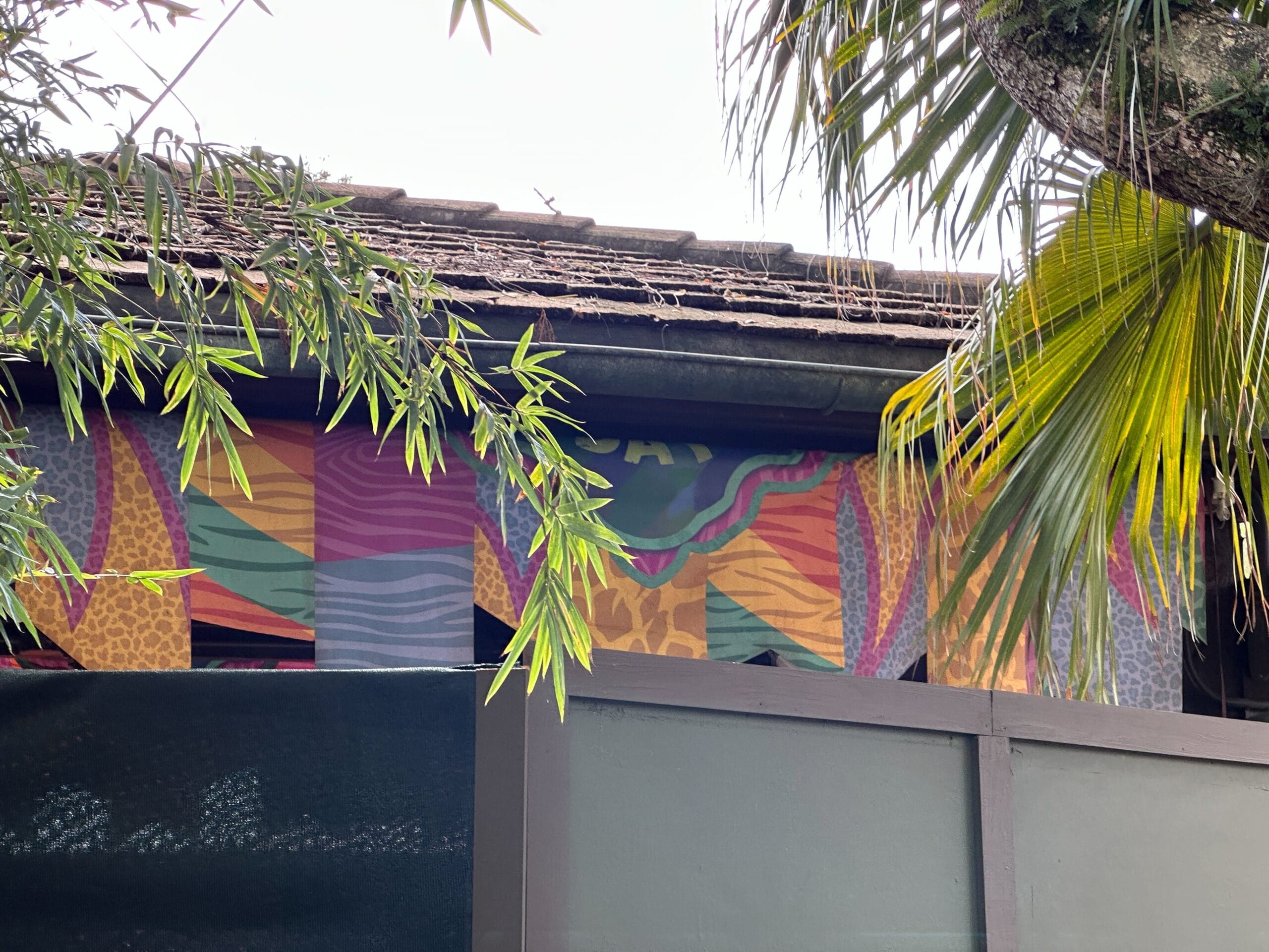 Brightly patterned fabric draped above a rooftop, green palm leaves in front, reminiscent of Adventureland at Disney Parks.