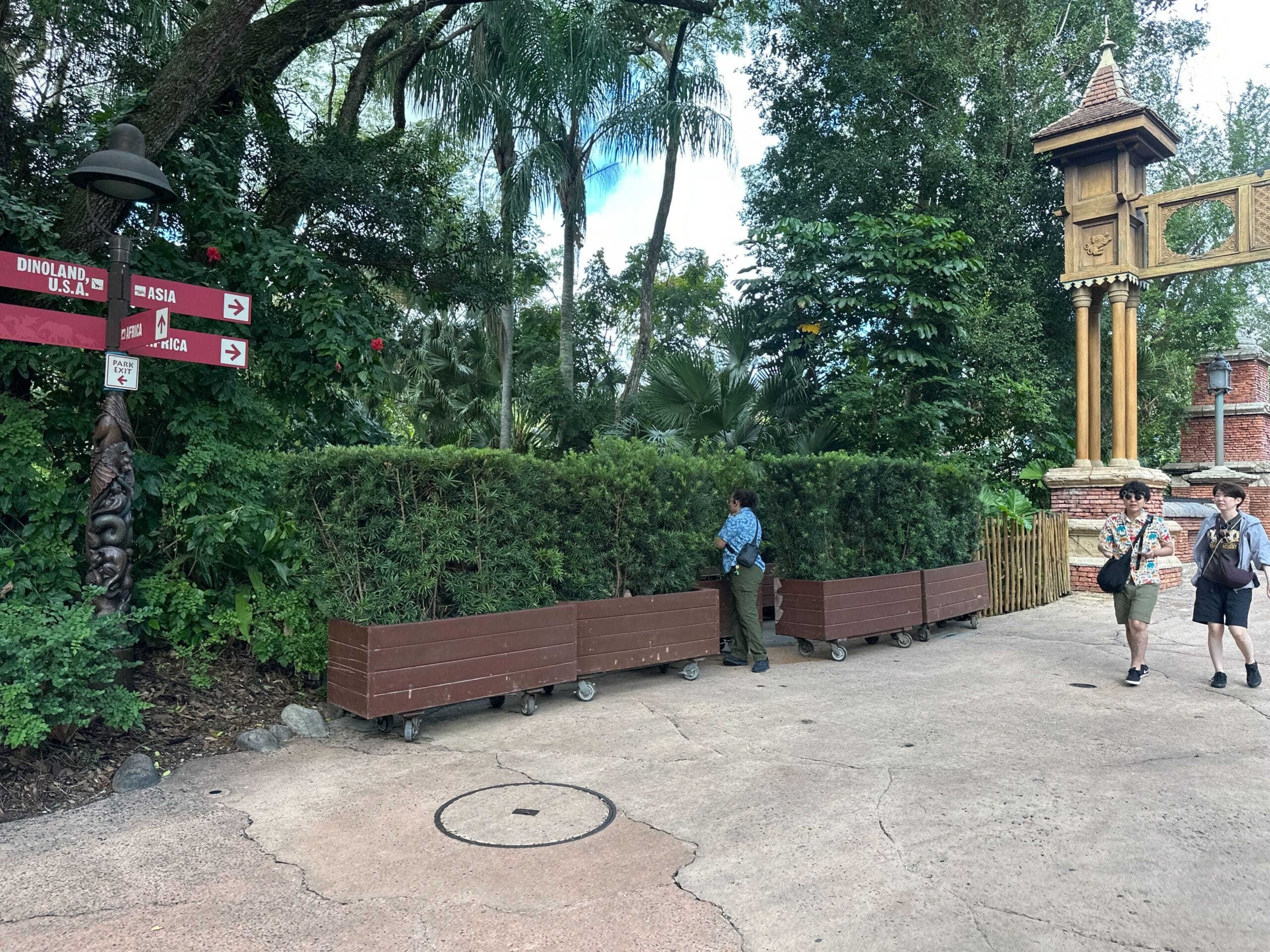 Guests stroll past a cast member trimming hedges near Zootopia signs amid lush Disney park landscaping, no castle in sight.