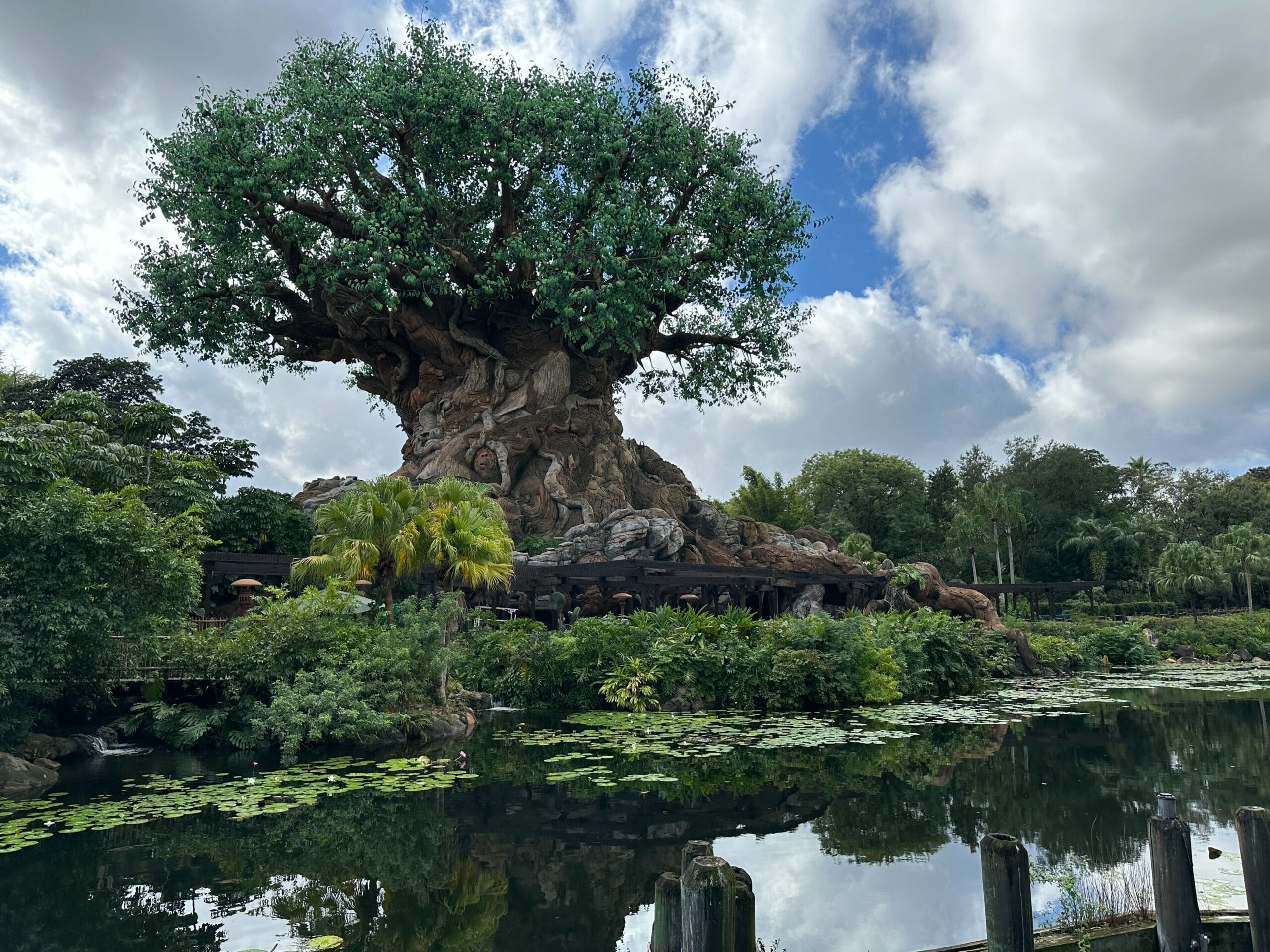 A massive artificial tree with animal carvings by a pond evokes Zootopia, creating a Disney park feel beneath a partly cloudy sky.