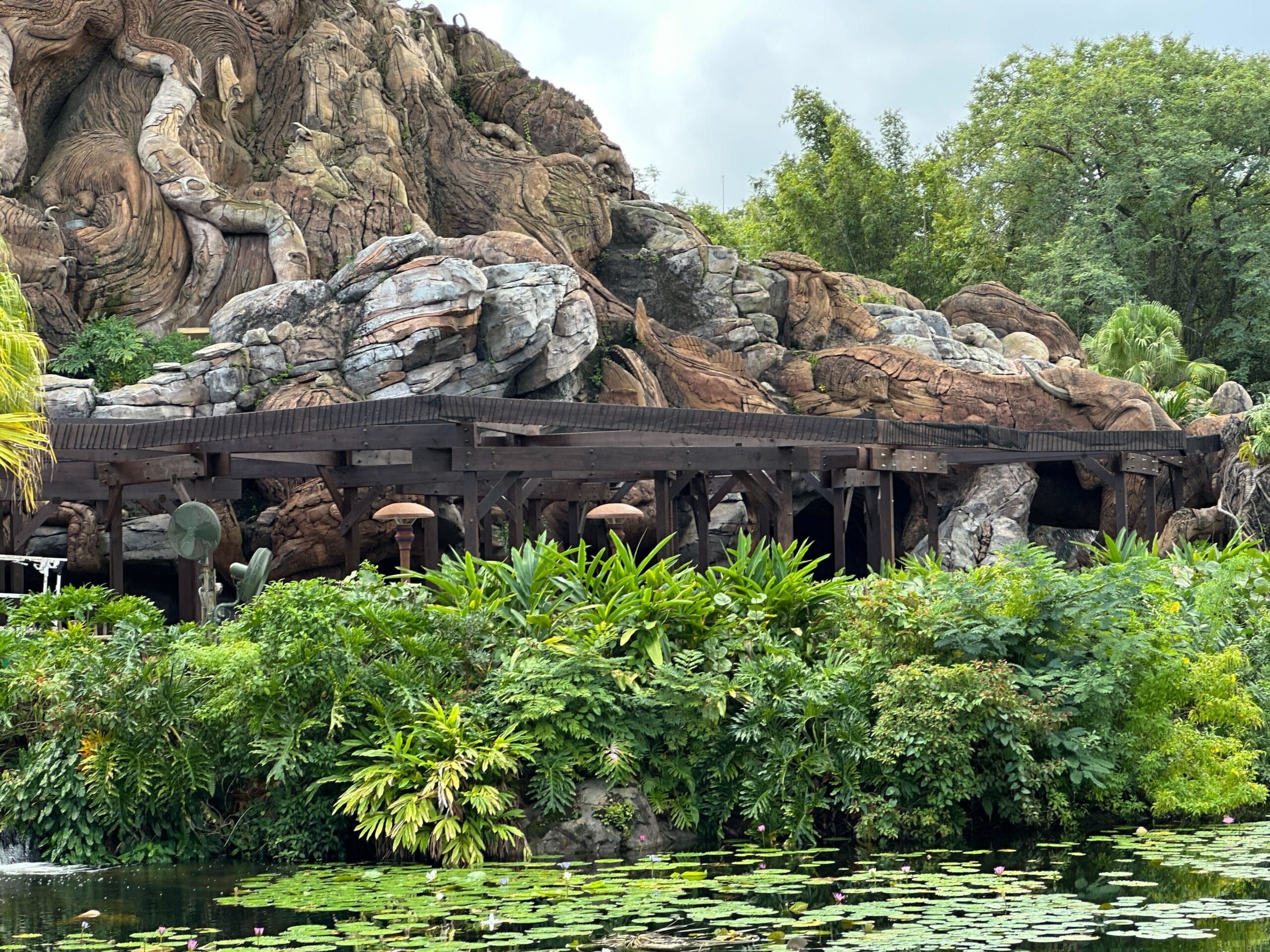 Verdant landscaping and reflective pond frame Zootopia mountain, cloudy skies above; "Better Zoogether" on the Disney park boardwalk.