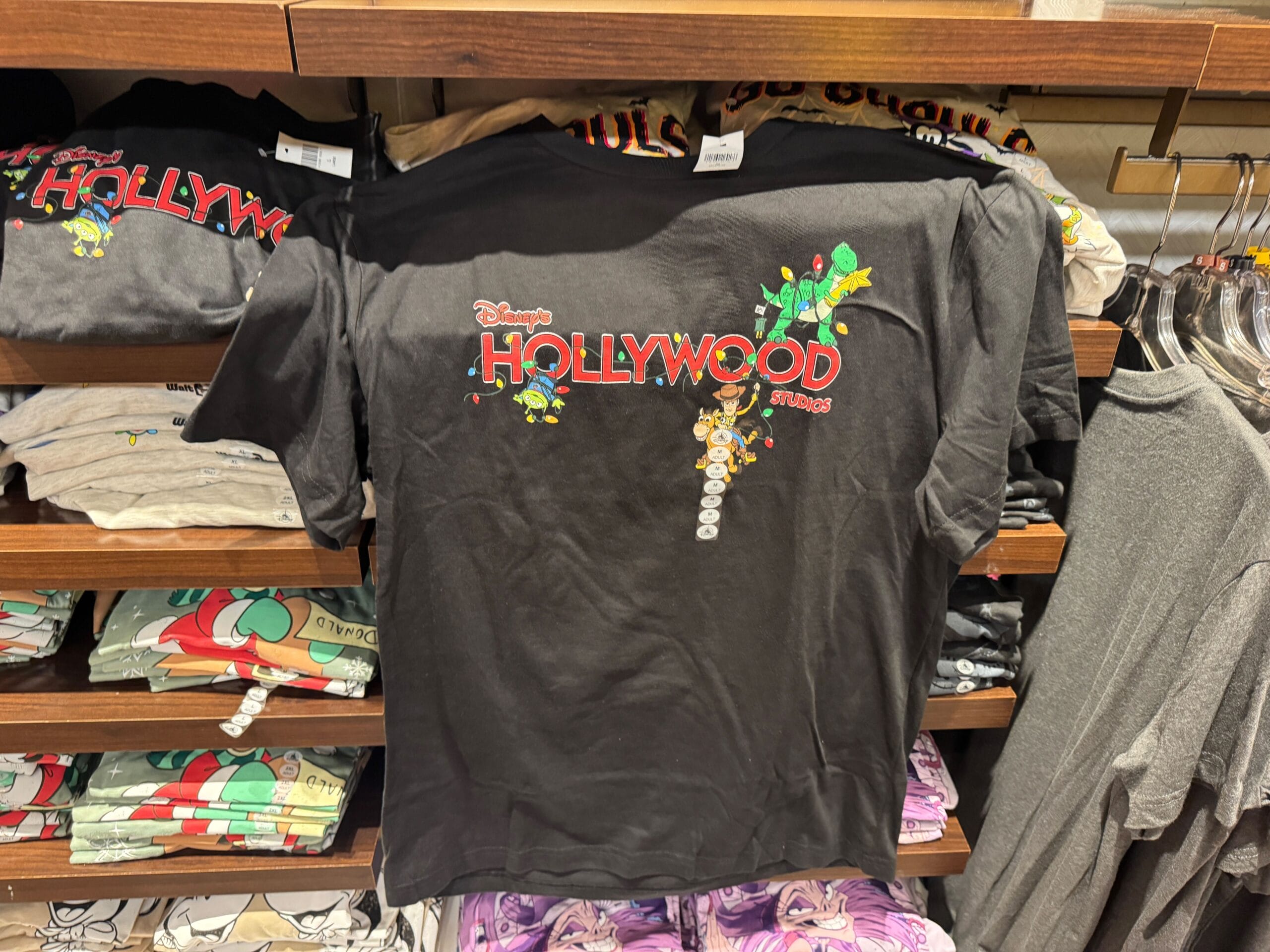 Black T-shirt with Disney's Hollywood Studios design, cartoon characters, and palm trees on a shelf; no castle like Disney parks' icons.