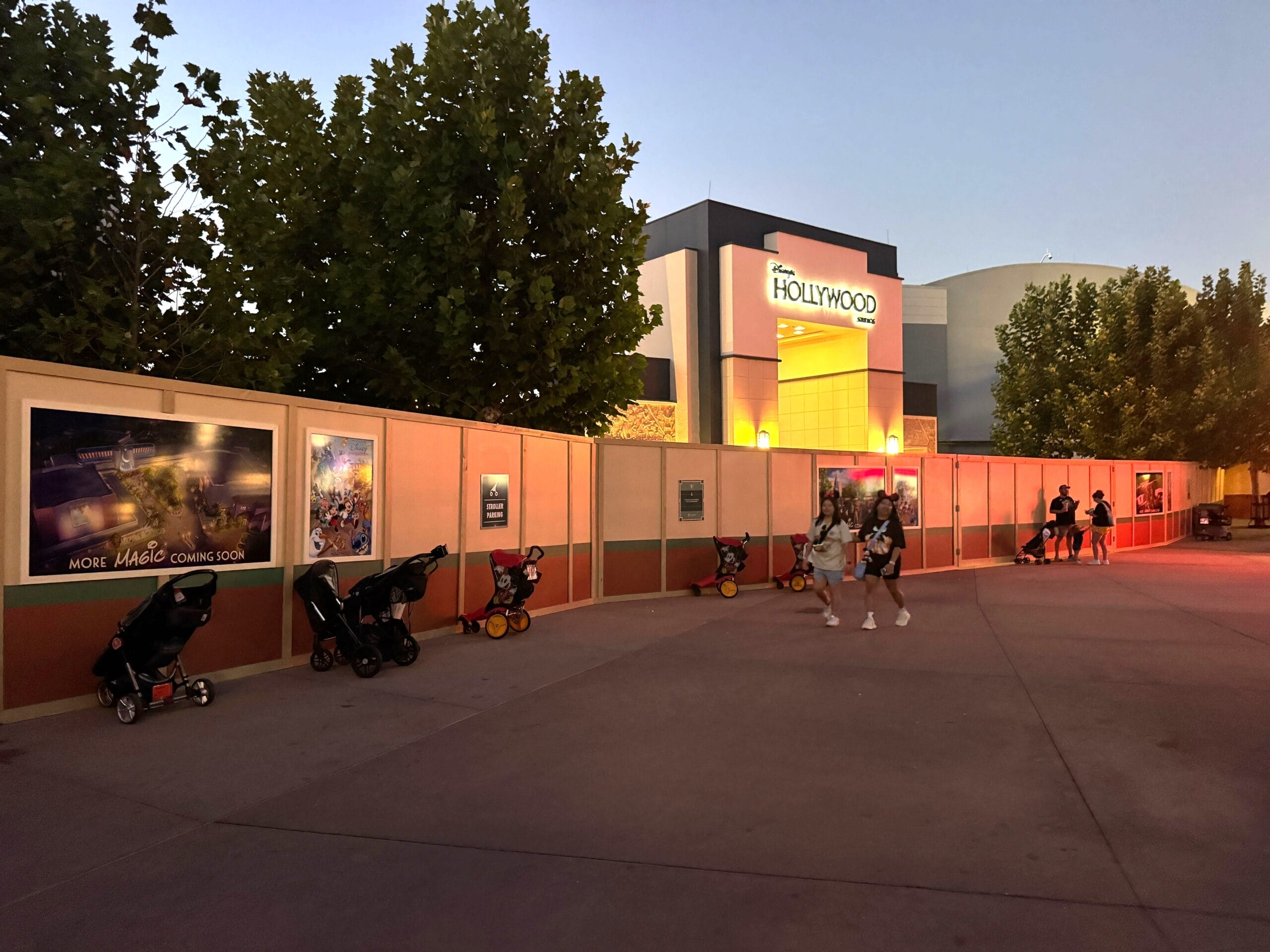 Empty strollers lined beside construction walls near a Hollywood-themed building at dusk—Disney park update, no castle in view.