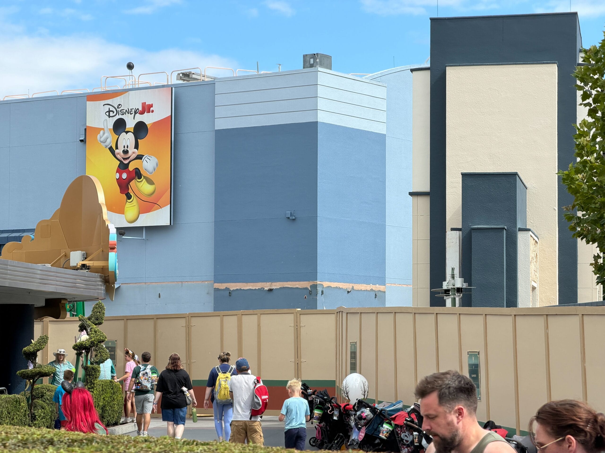 Guests stroll by construction walls surrounding the demolition of the former Animation Courtyard in Disney's Hollywood Studios. A Disney Junior Mickey Mouse sign hangs above on a blue building.