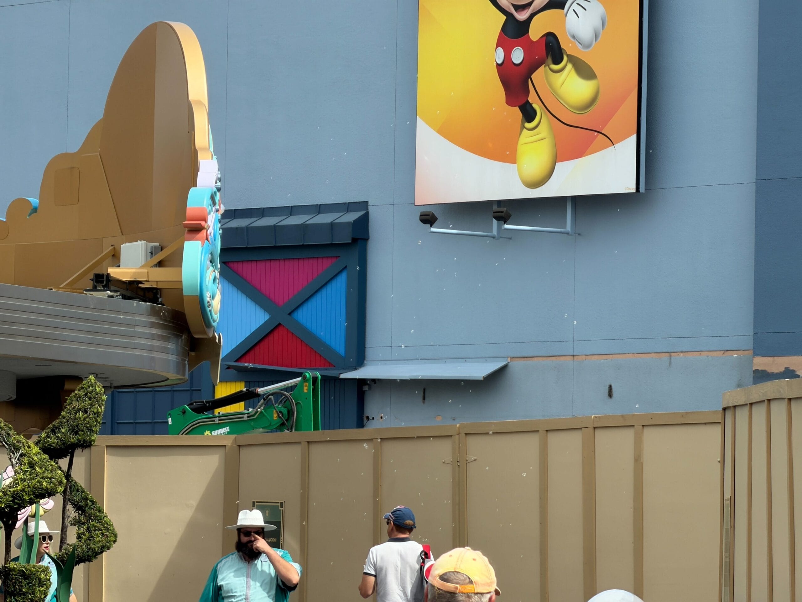 Guests gather by a tan construction wall at a Disney park, with a Mickey Mouse emblem above—no castle in view to compare.