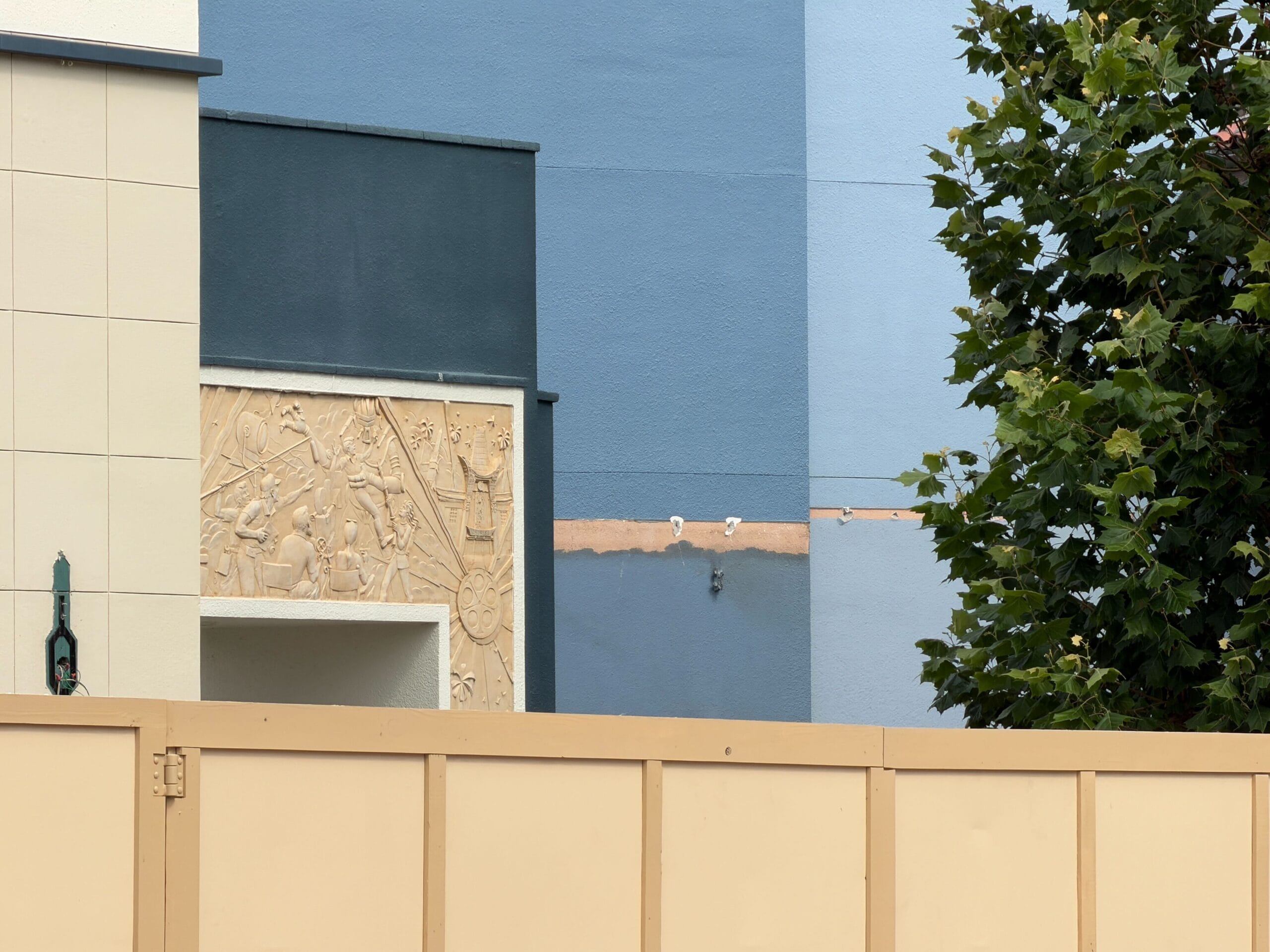 A beige construction fence in front of a blue building, tree, and decorative wall relief—typical of Disney park refurbishments.