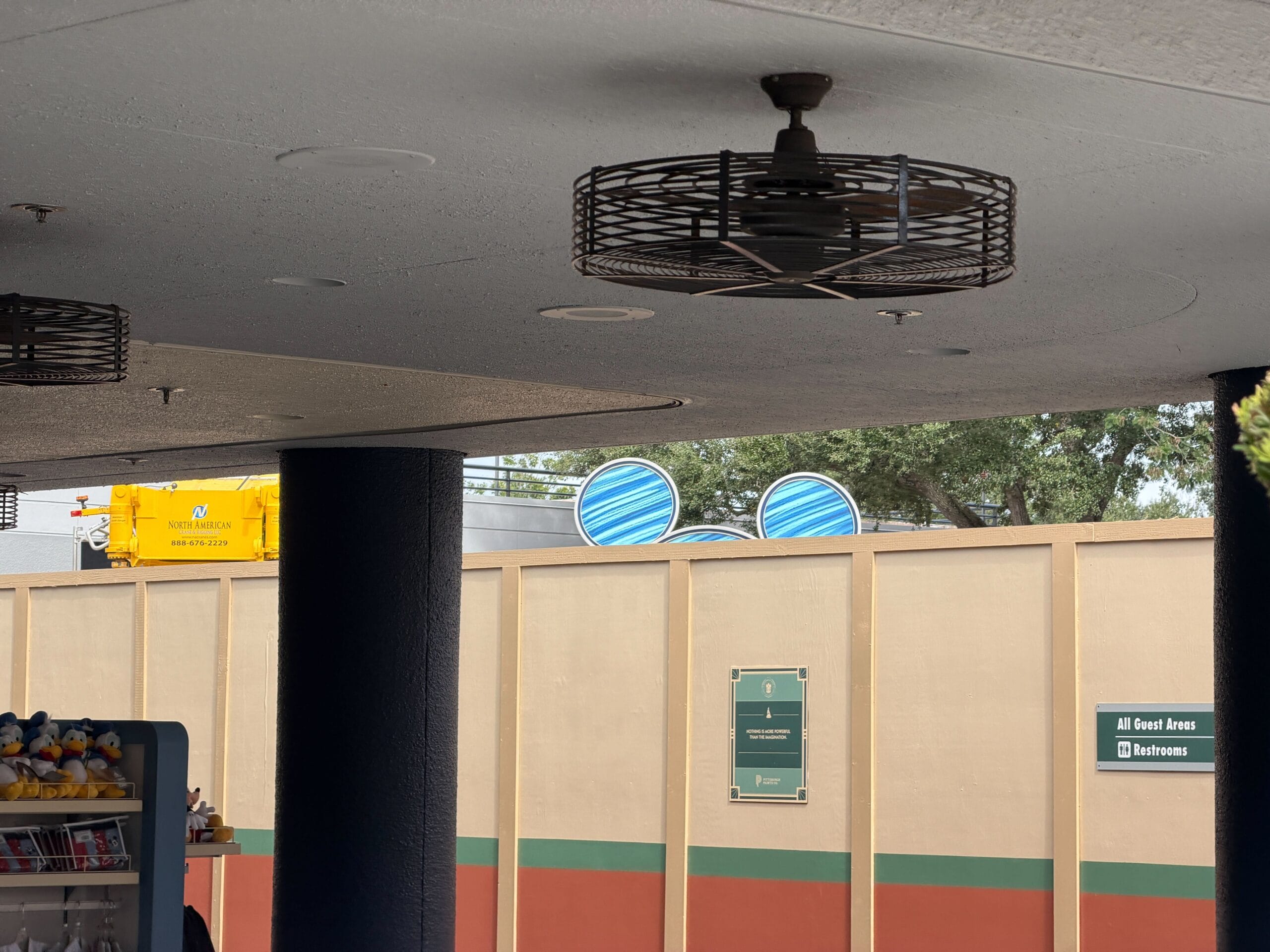 Two large blue spinning discs behind a tan construction wall in a Disney park; fans and merchandise shelves in the foreground.