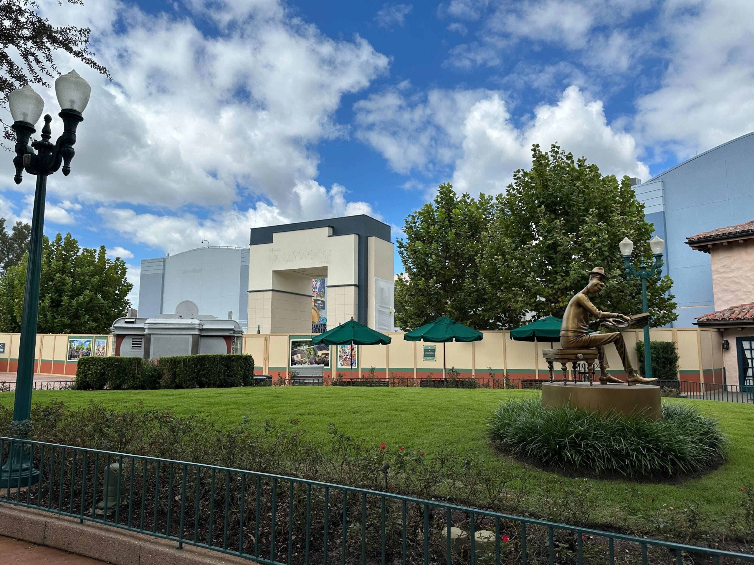 A bronze statue of a man sits on a bench at Disney's Hollywood Studios park, trees and umbrellas around, blue skies above.