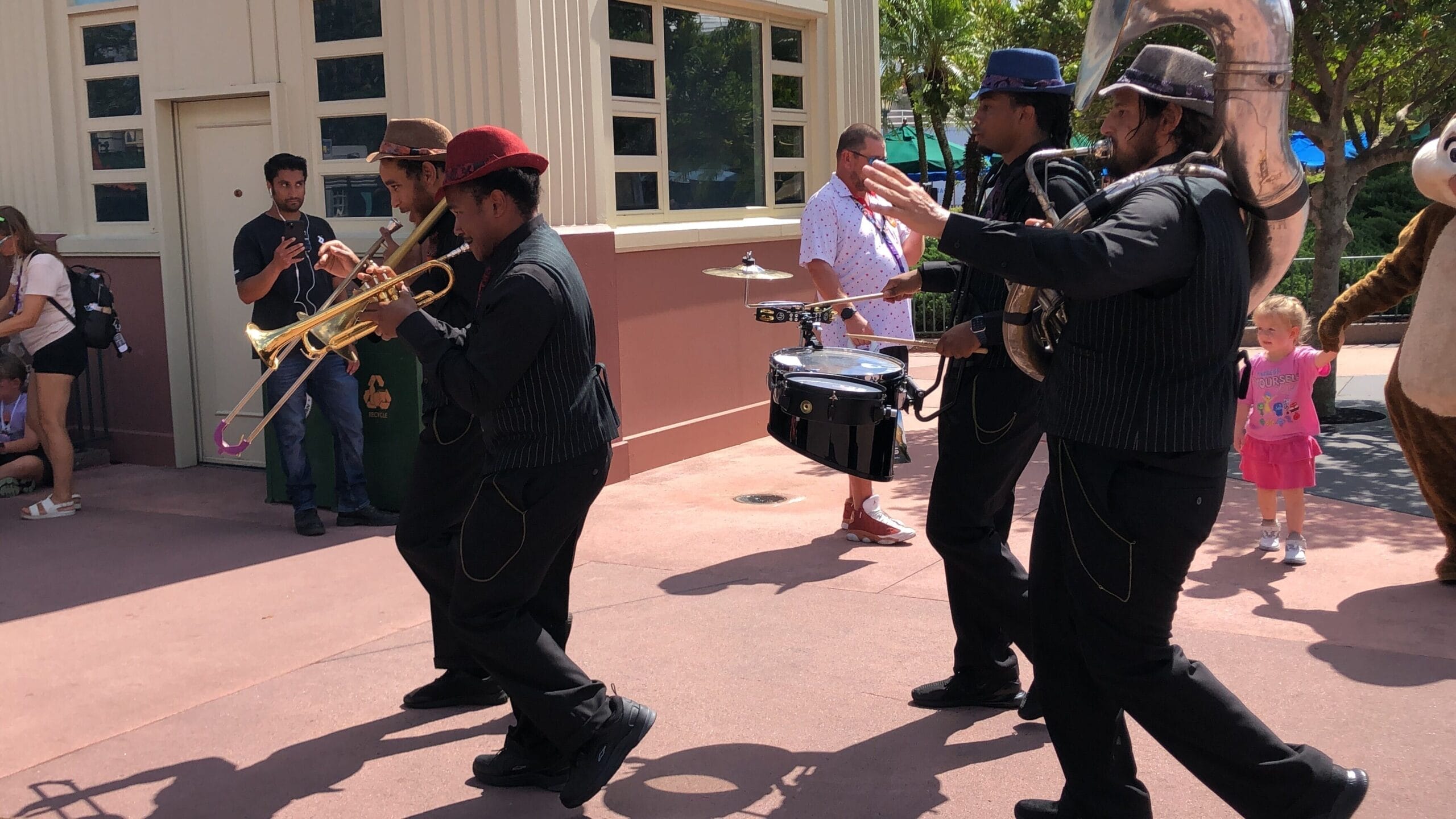 Four musicians from the HollyGroove Swinging Band play instruments outdoors at Hollywood Studios, with bystanders nearby.