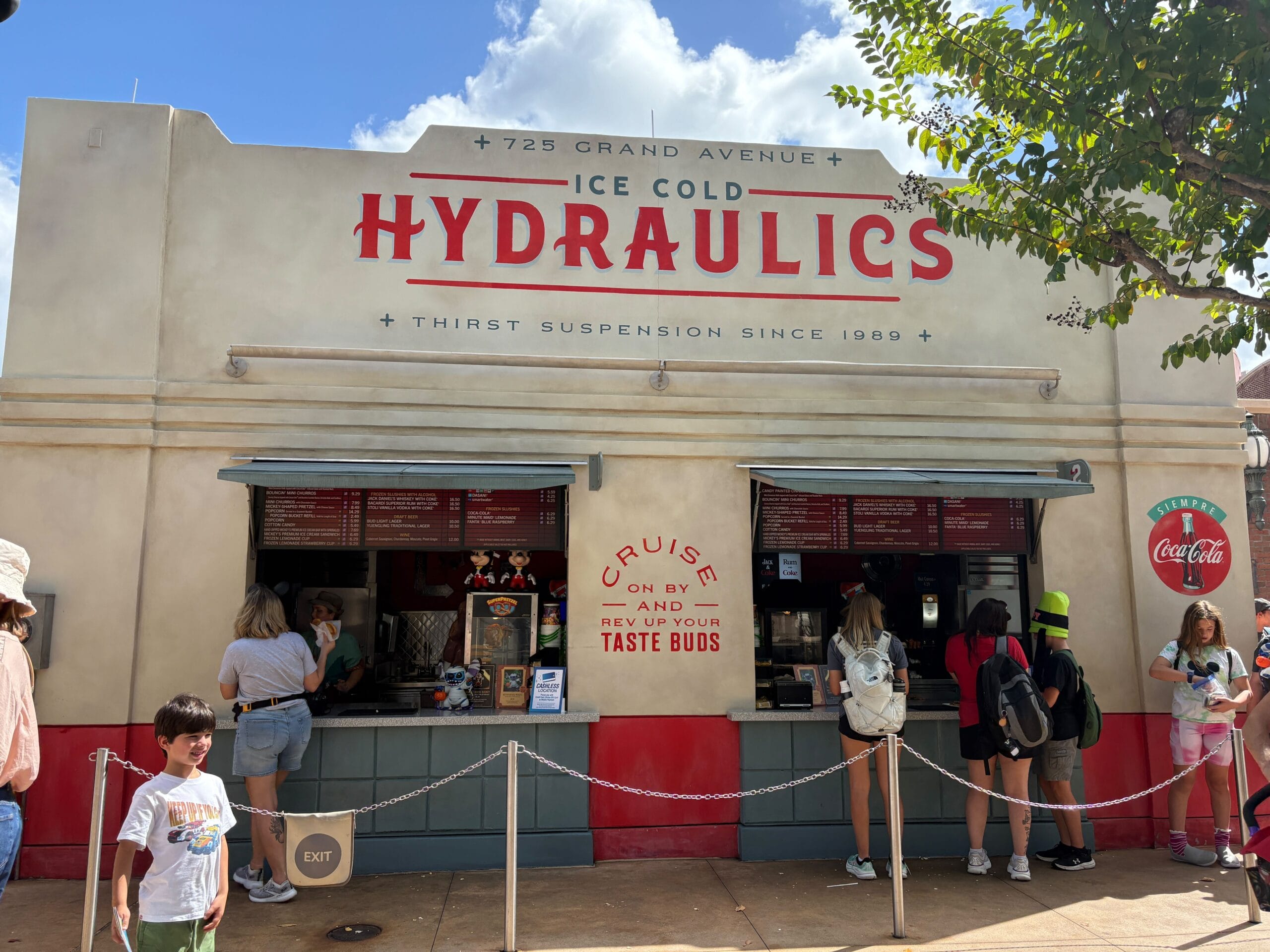 Guests line up at a retro Walt Disney World snack cart with a Coca-Cola sign, enjoying a sunny day in the park.