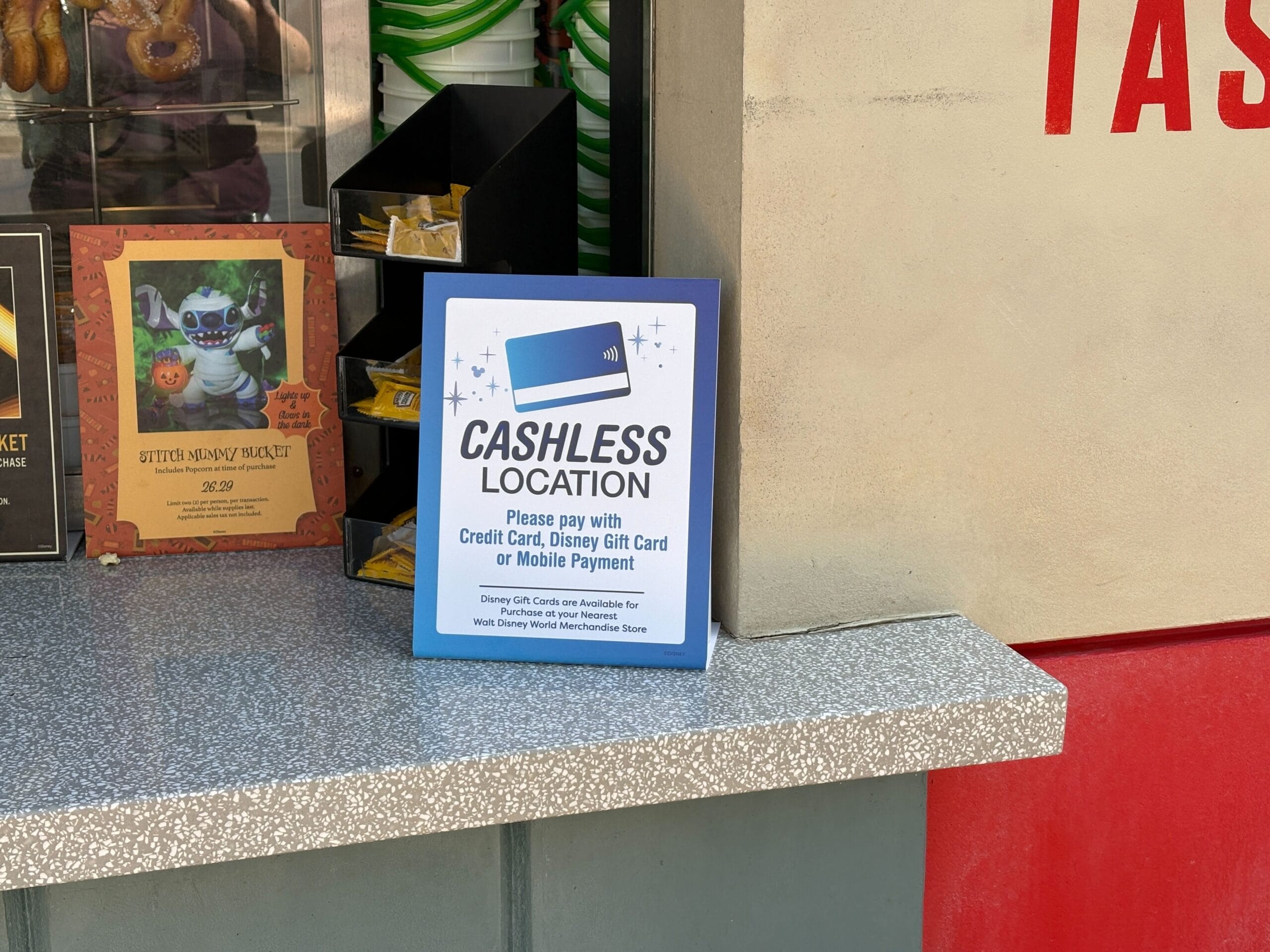 A Walt Disney World snack cart sign says "Cashless Location," instructing guests to use cards or mobile pay—no castle visible.