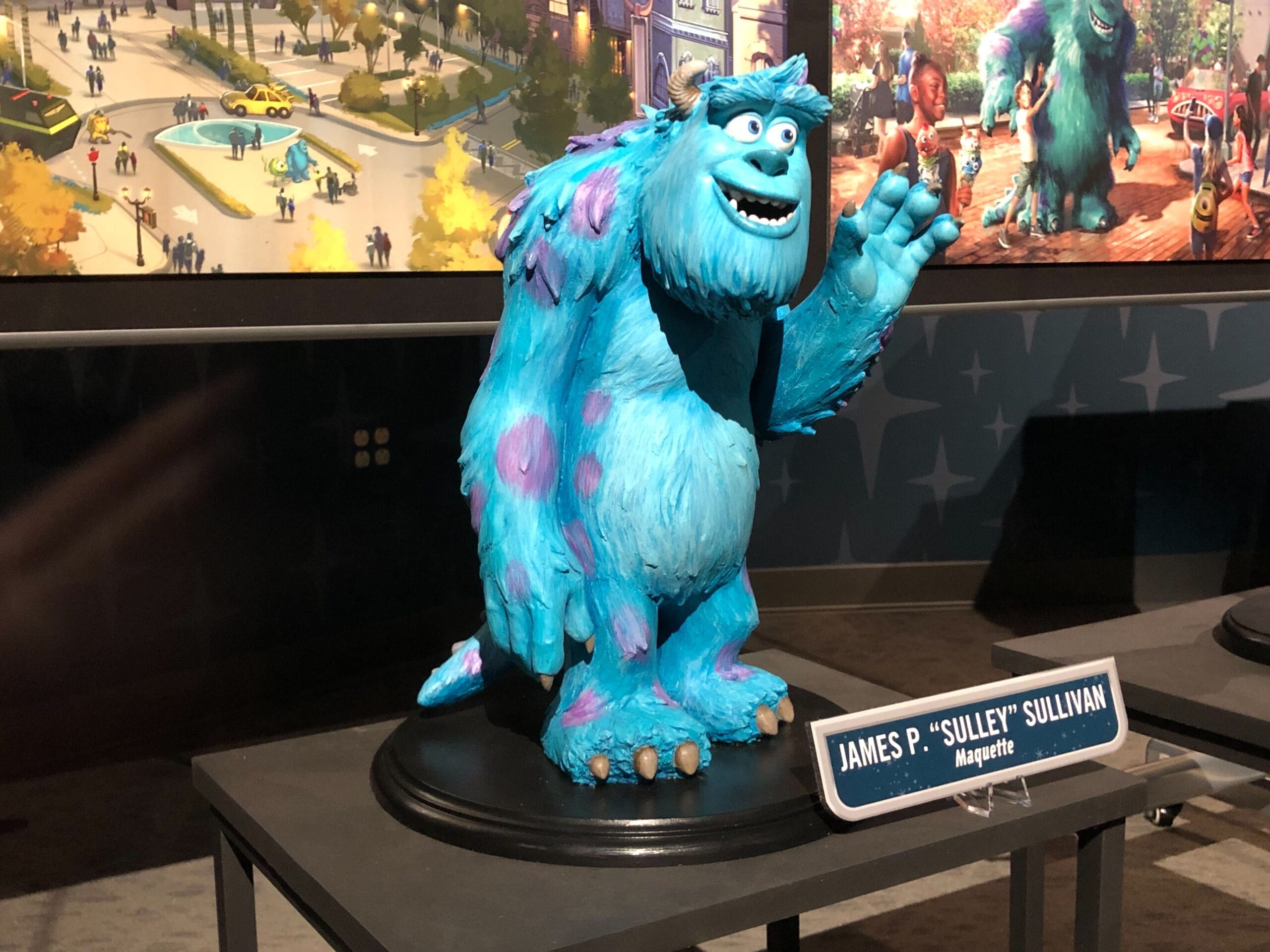 A Sulley statue from Monsters, Inc. stands on a nameplate before a bright Monsters Inc. Coaster backdrop, reminiscent of Disney parks.