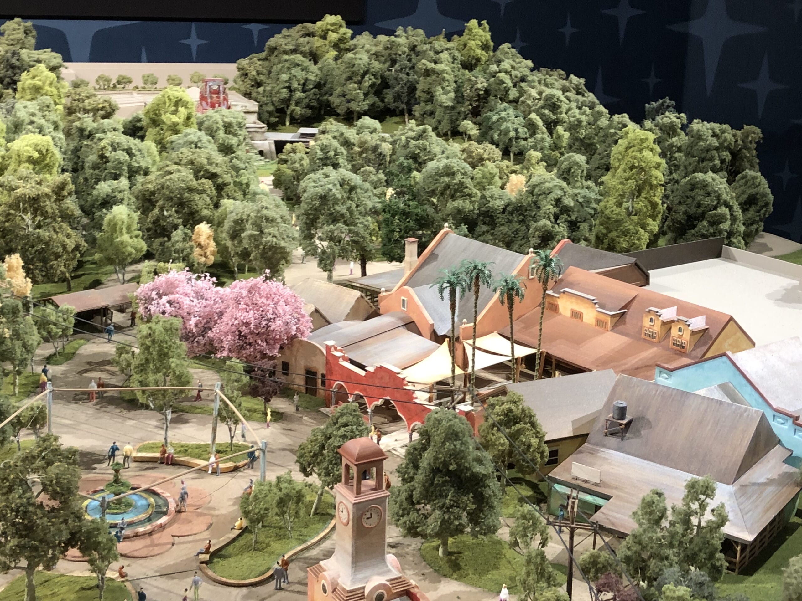 A detailed theme park model highlights Cars Land buildings, a tower, greenery, and miniature guests near a small fountain—no castle present.