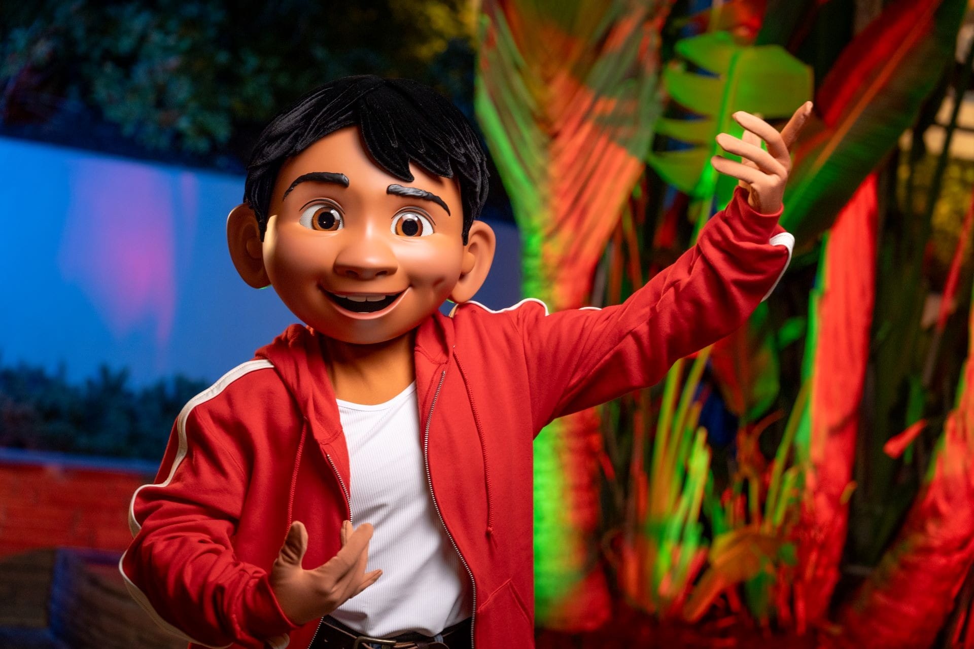 Animated character in a red jacket poses at Disney Jollywood Nights 2025, surrounded by vivid lights and lush tropical plants—no castle visible.