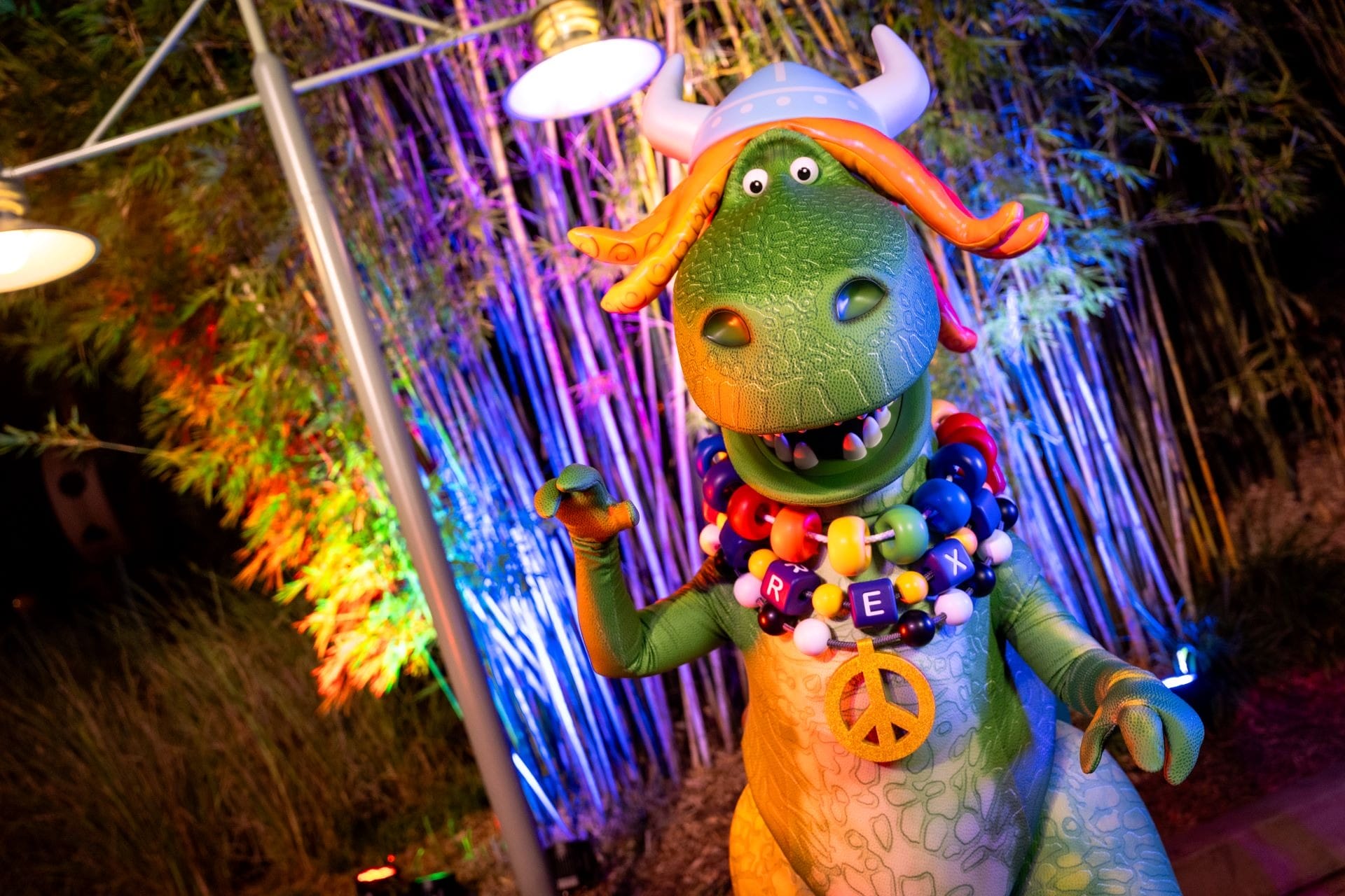At a Disney park, Partysaurus Rex the green dino wears a party hat, balloon necklace, and peace sign with dazzling colorful lights.