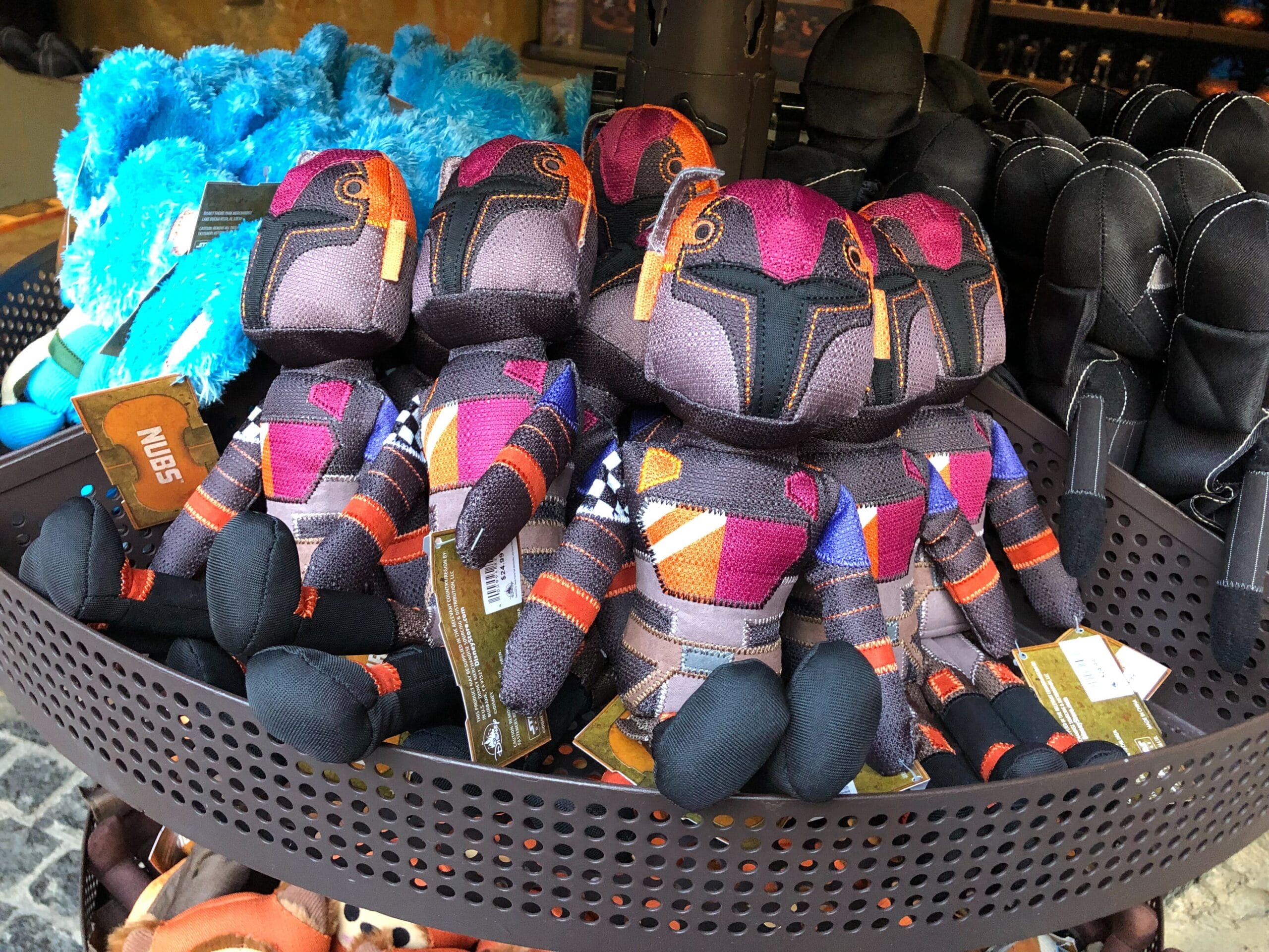 Plush Sabine Wren Doll in purple and orange armor, displayed with other plushes at Star Wars Galaxy's Edge in a Disney park setting.