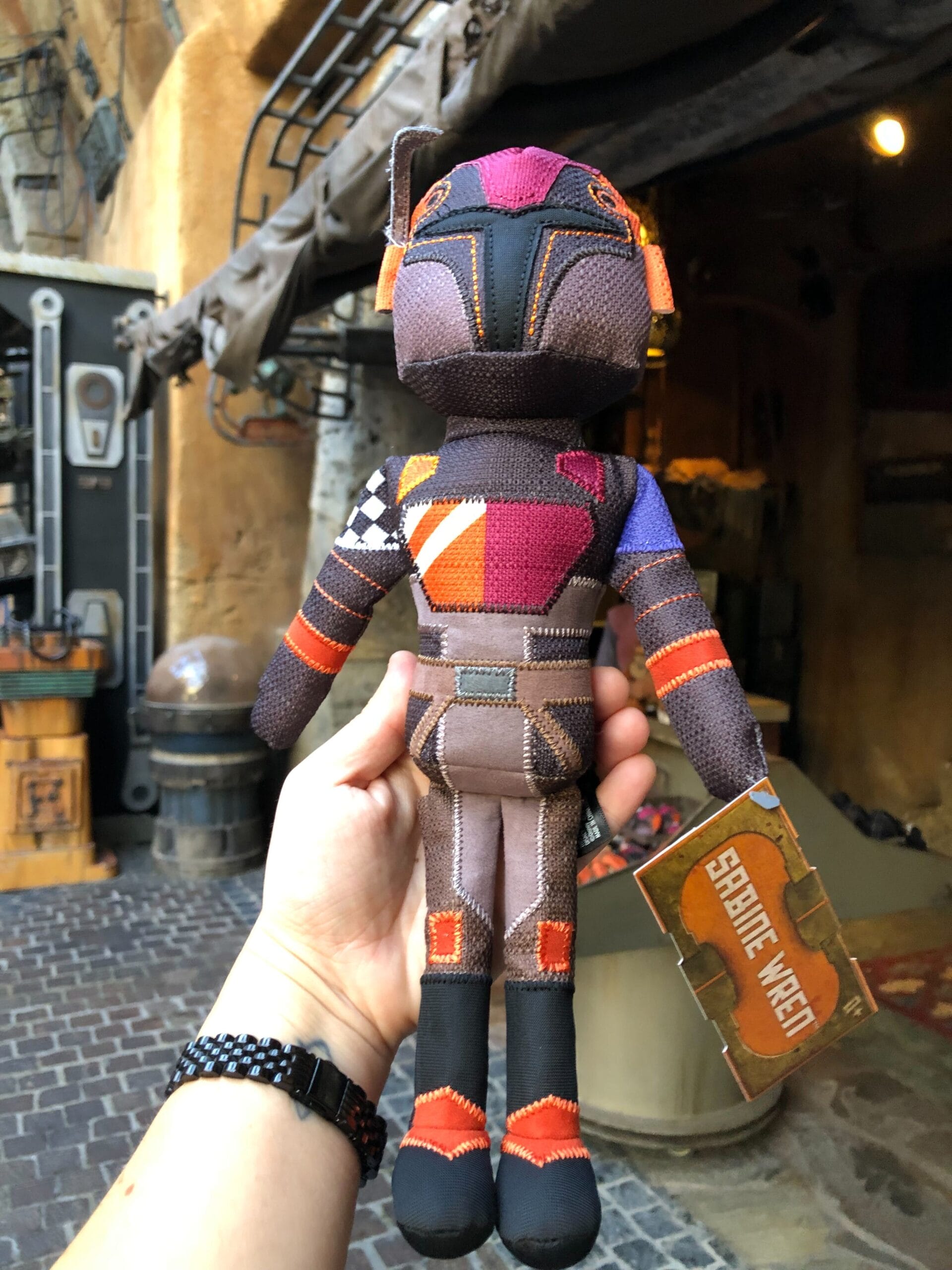 A hand holds a Sabine Wren Doll from Star Wars at Galaxy's Edge in Disney Parks, with rustic market decor typical of Batuu markets.