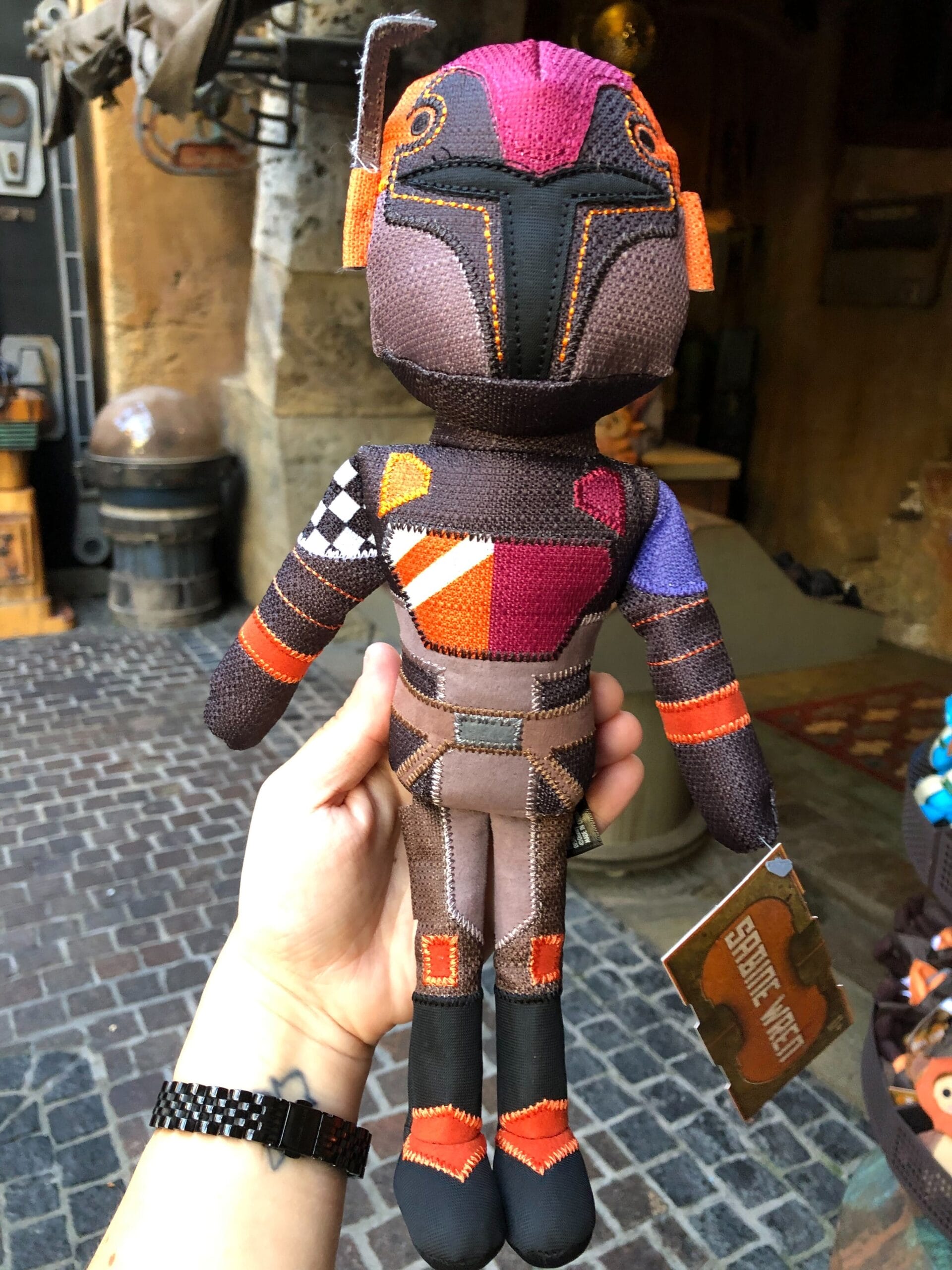 A hand displays a bright Sabine Wren doll with tag at Disney's Star Wars Galaxy's Edge, no castle in view to compare to any parks.