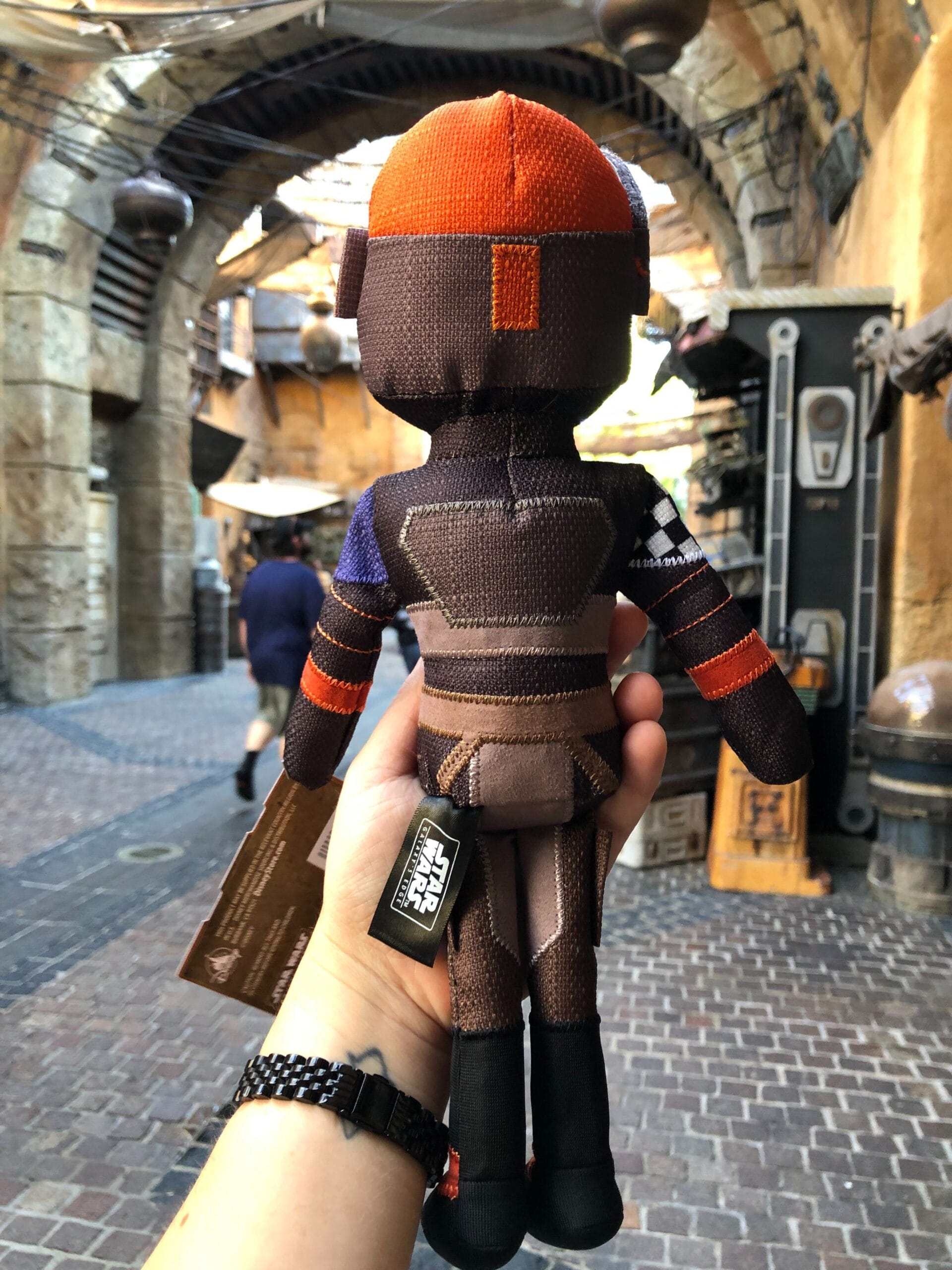 A hand displays a Sabine Wren Doll at Star Wars Galaxy’s Edge, framed by Batuu’s stone arches—no castle like Disney’s here.