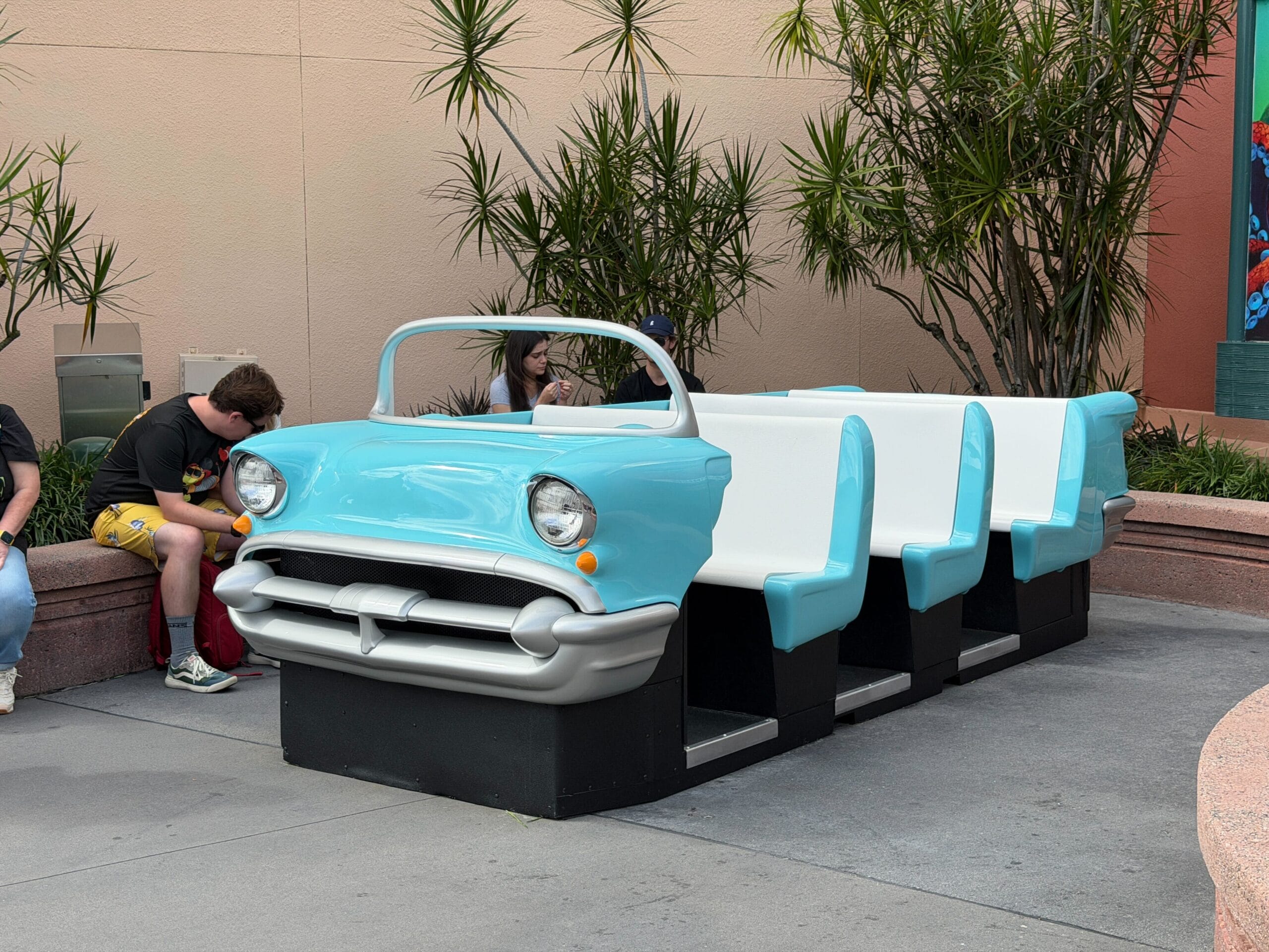 A whimsical bench styled like a vintage blue car with white seats, outdoors near greenery and guests—reminiscent of Disney park decor.