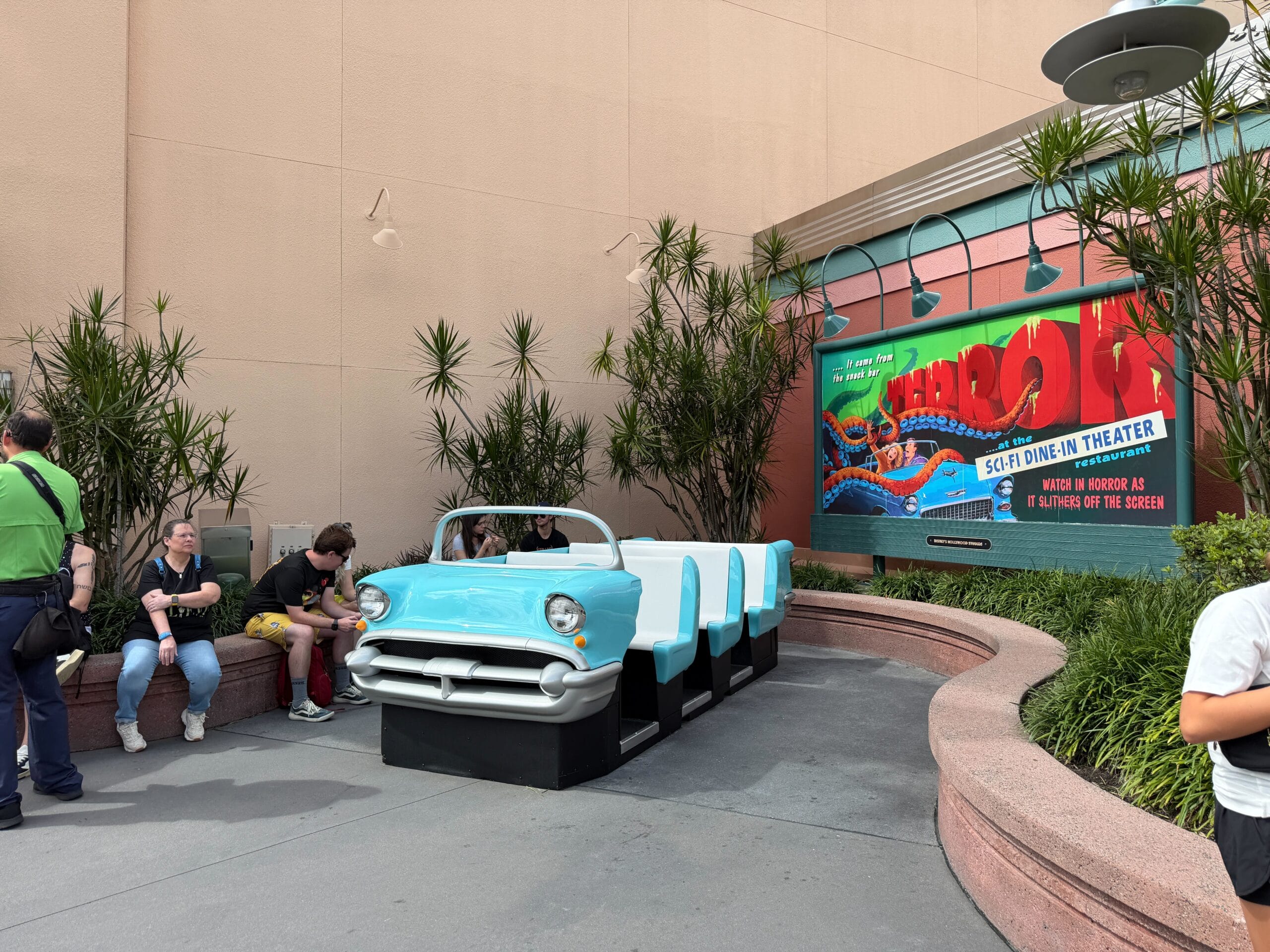 Guests relax by a blue car-shaped booth under the vintage "Sci-Fi Dine-In Theater" sign, unlike any Disney park castle scene.