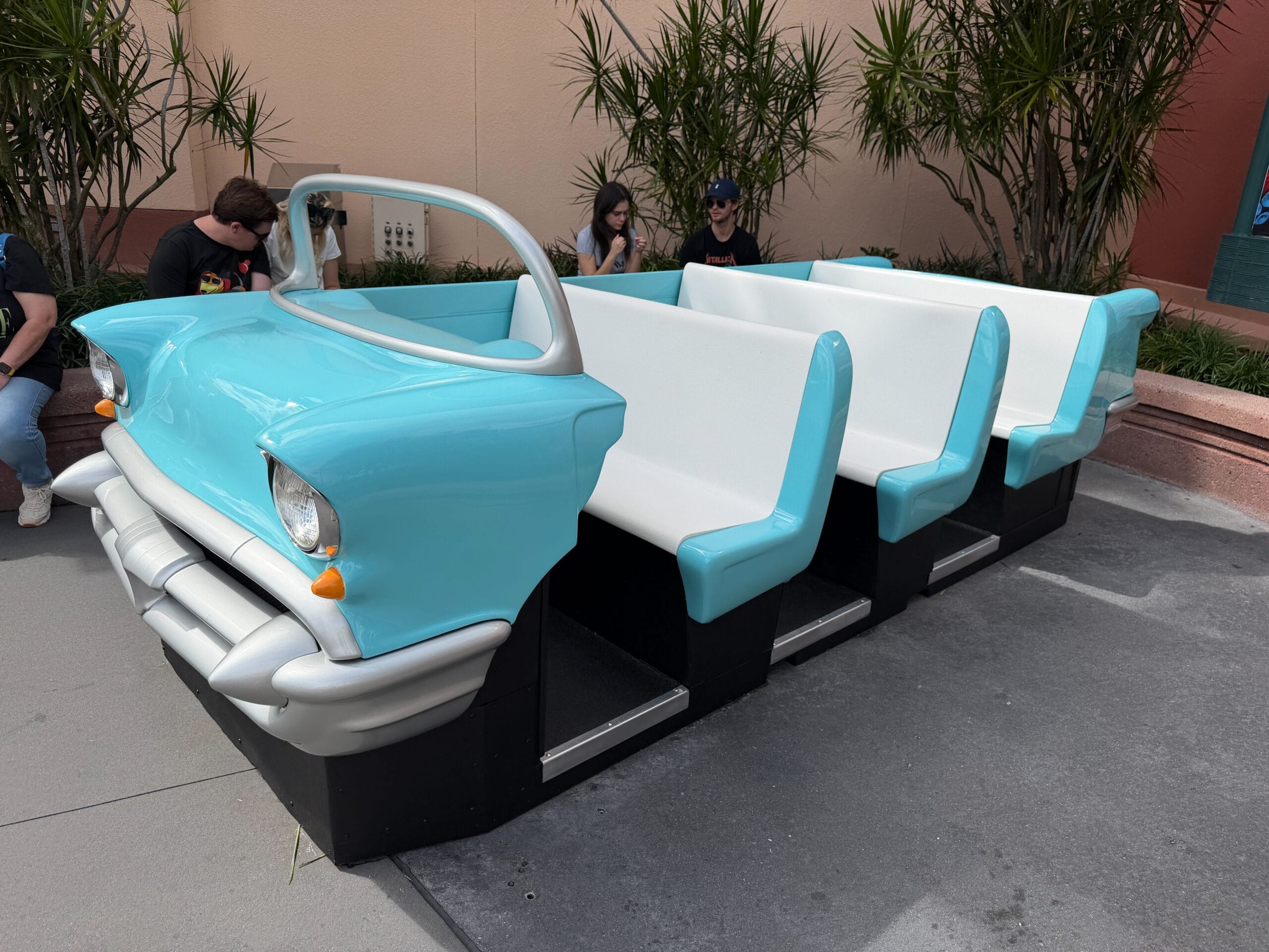 A light blue and white bench shaped like a vintage car, outside near greenery and guests, evokes whimsical Disney park design.