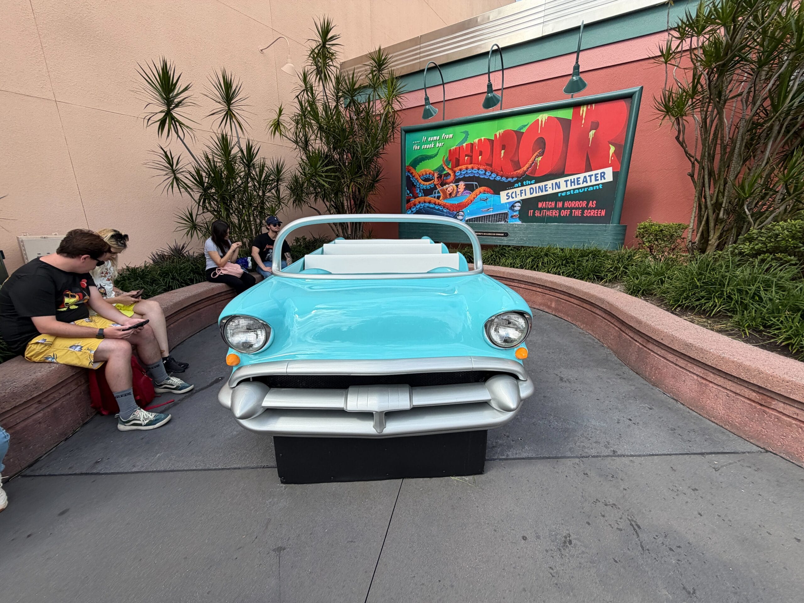 A light blue vintage car photo op sits before a retro movie poster, with guests relaxing nearby—classic Disney park ambiance.